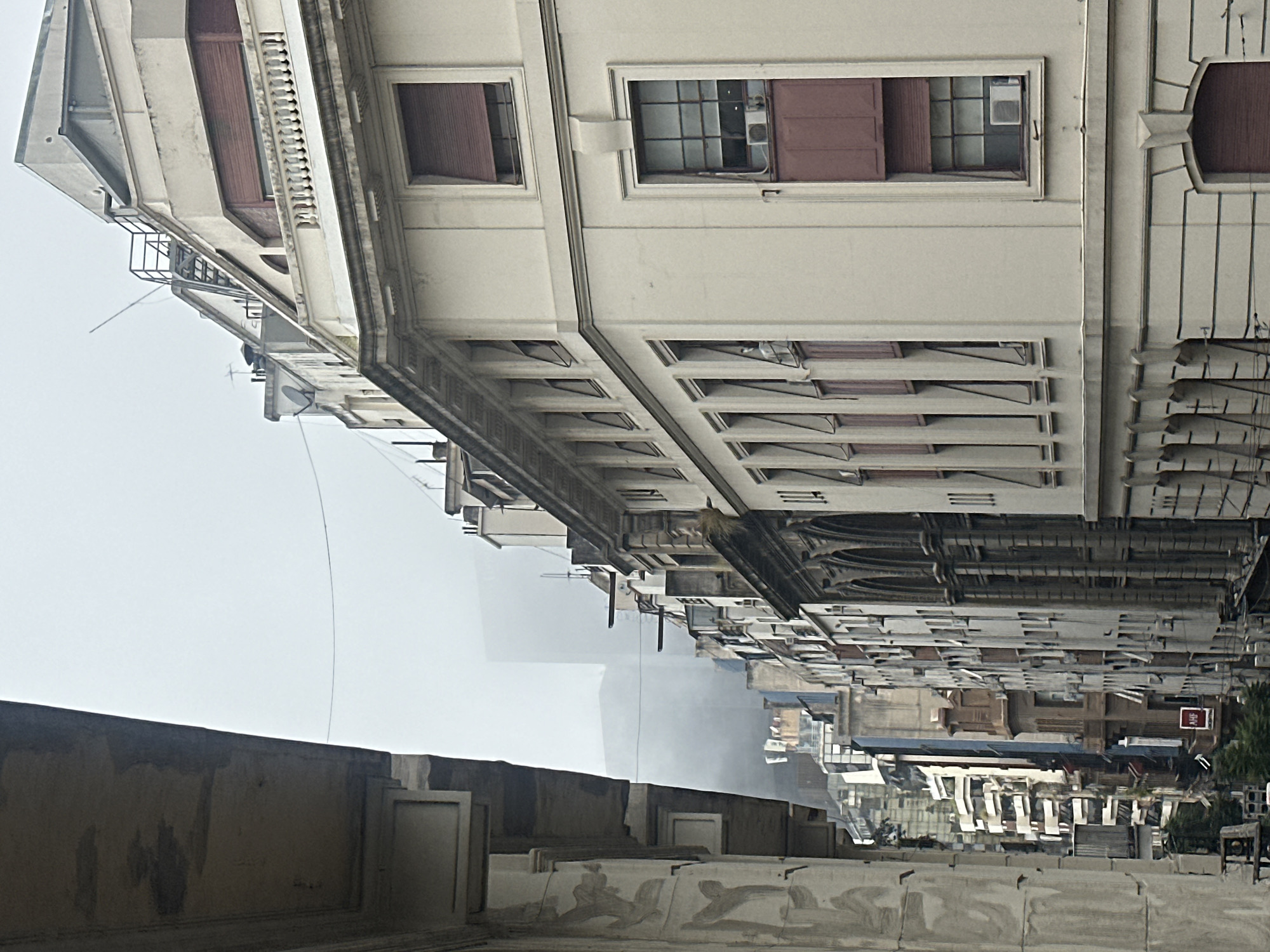 The image shows a street in Buenos Aires, Argentina, looking downward from an elevated position. The architecture is characterized by tall, narrow buildings with ornate facades, many featuring window shutters, and detailed cornices. Several buildings show signs of age and weathering. A visible portion of a wall or ledge, constructed of weathered concrete, occupies the foreground. In the distant center, a line of white buildings fades into the overcast sky. Other buildings are visible, with the street ending far away with a bench to the right and a red sign to the right of the shot.