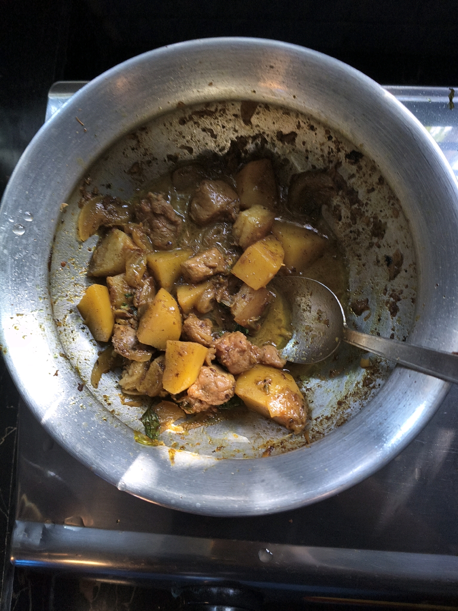 The image shows a close-up, overhead view of a round, shallow, metallic pot filled with a stew-like dish. The dish contains chunks of meat and cubed potatoes in a dark, oily sauce. A metal ladle with a wide, shallow bowl rests in the pot, partially submerged in the food. The pot is placed on a dark, reflective surface, possibly a stovetop. The background is dark and out of focus. There are no people or animals visible in the image. The overall impression is of a home-cooked meal. The image's origin in Rangamati, Bangladesh is not discernible from the visual content itself, which focuses exclusively on the food and the cooking vessel.