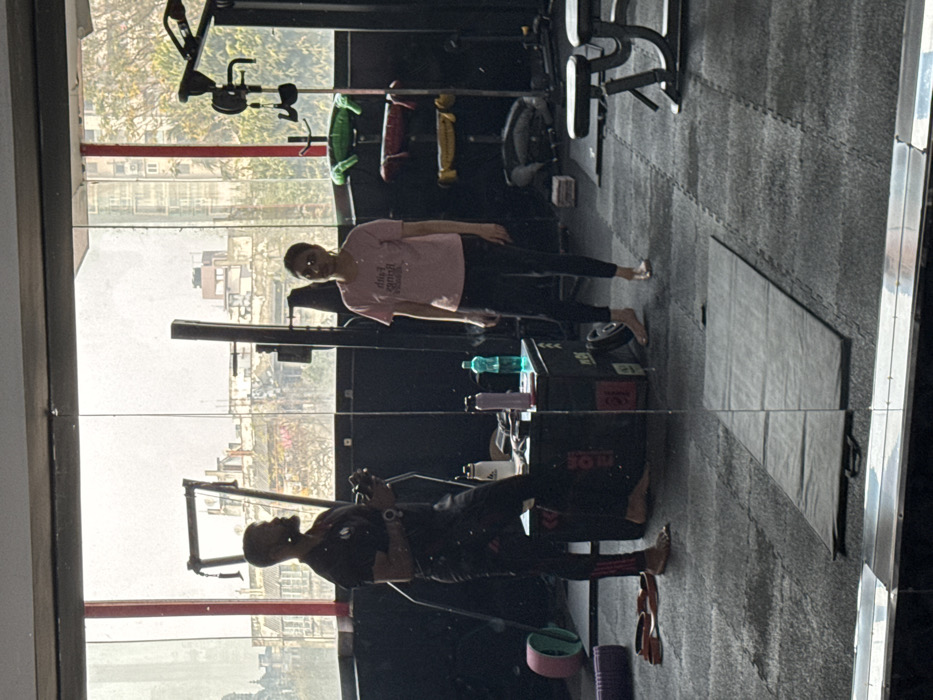 The image shows a gym's interior through a glass panel, reflecting the scene. A woman in a pink shirt and black leggings stands in the gym, and a man is working out. Gym equipment, including a cycling machine and weight racks, are visible in the background. A cityscape can be seen through the gym windows. The reflection creates a mirror image of the gym space.