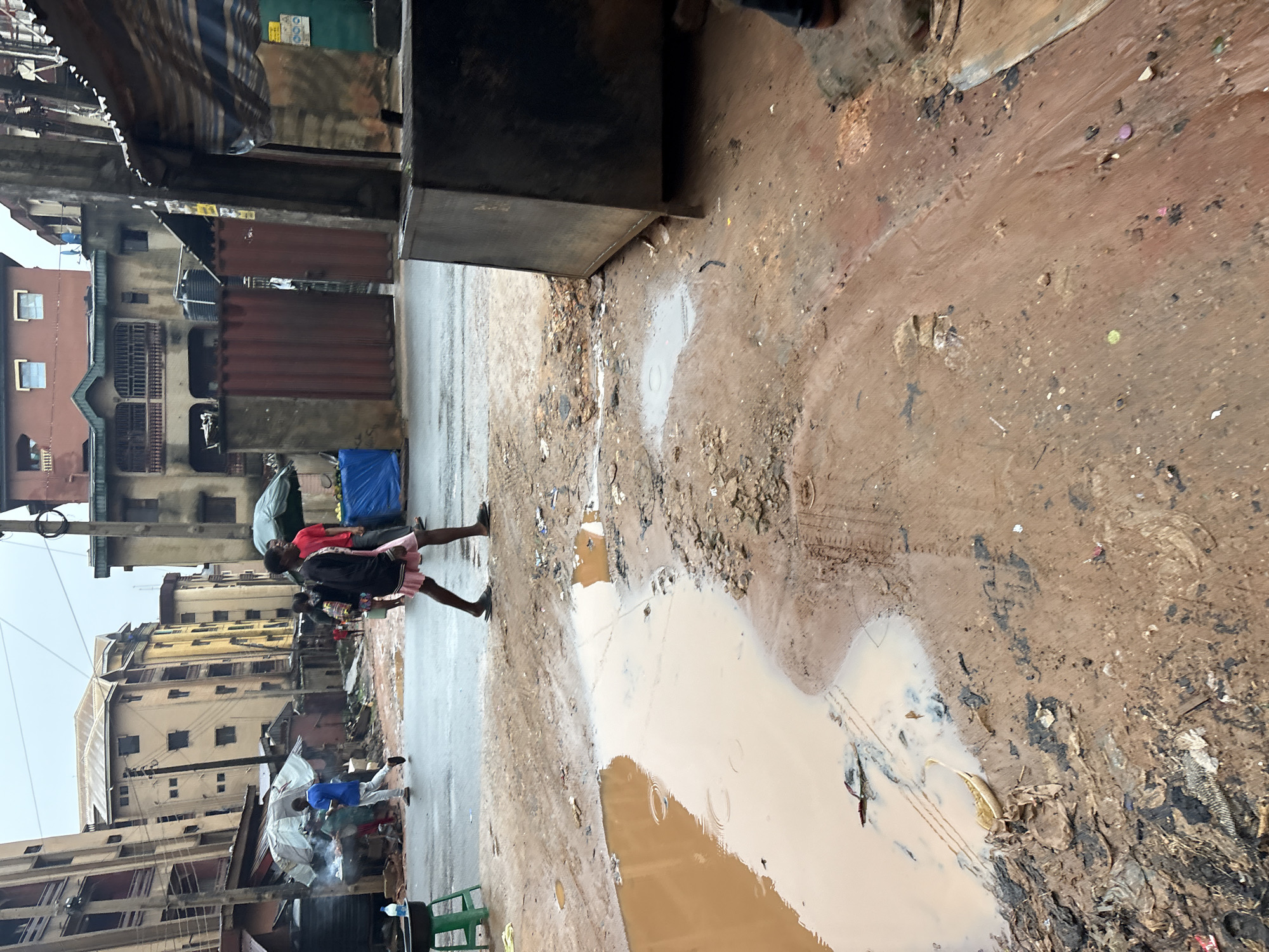 The image depicts a street scene in Onitsha, Nigeria, characterized by its wet and muddy conditions, likely due to recent rainfall. The ground is covered in brown mud and debris, with a large puddle dominating the foreground. Several people are visible walking or standing. In the mid-ground, a person in a red shirt and a person in a black jacket and pink shorts are walking on the wet street. Further back, a person in a blue shirt is walking. The background features multi-story buildings with balconies and windows, typical of urban architecture. There are also market stalls or makeshift shelters with awnings, suggesting a commercial area. The overall atmosphere is one of a bustling, yet weathered, urban environment with evidence of everyday life and challenging conditions.