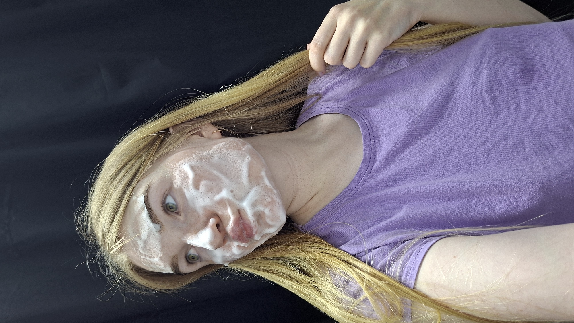 A fair-skinned young woman with long blonde hair is lying down on a black surface, possibly fabric. She is wearing a purple t-shirt. Her face is covered in white foam, possibly from a facial cleanser. Her eyes are open, and she is looking towards the viewer. One of her hands is near her hair.
There is no indication of location in Chaikovsky, Russia, or any other geographical location. The scene is a close-up of a person, and the background is a dark, indistinct surface.
