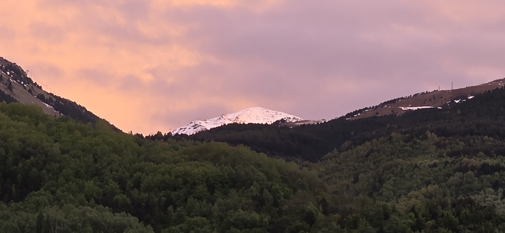 The image depicts a mountainous landscape at dusk. In the foreground, dense green forests cover the slopes of lower hills. Behind these, a larger, snow-capped mountain is visible with the sun reflecting off of its summit. To the left and right of the main mountain, additional tree-covered hills rise, one having ski-lift infrastructure. The sky is colored with hues of pink and orange, suggesting either sunrise or sunset. The scene appears to be located in the Pyrenees mountains, near the city of Benasque, Spain. Three radio towers are visible in the background.
