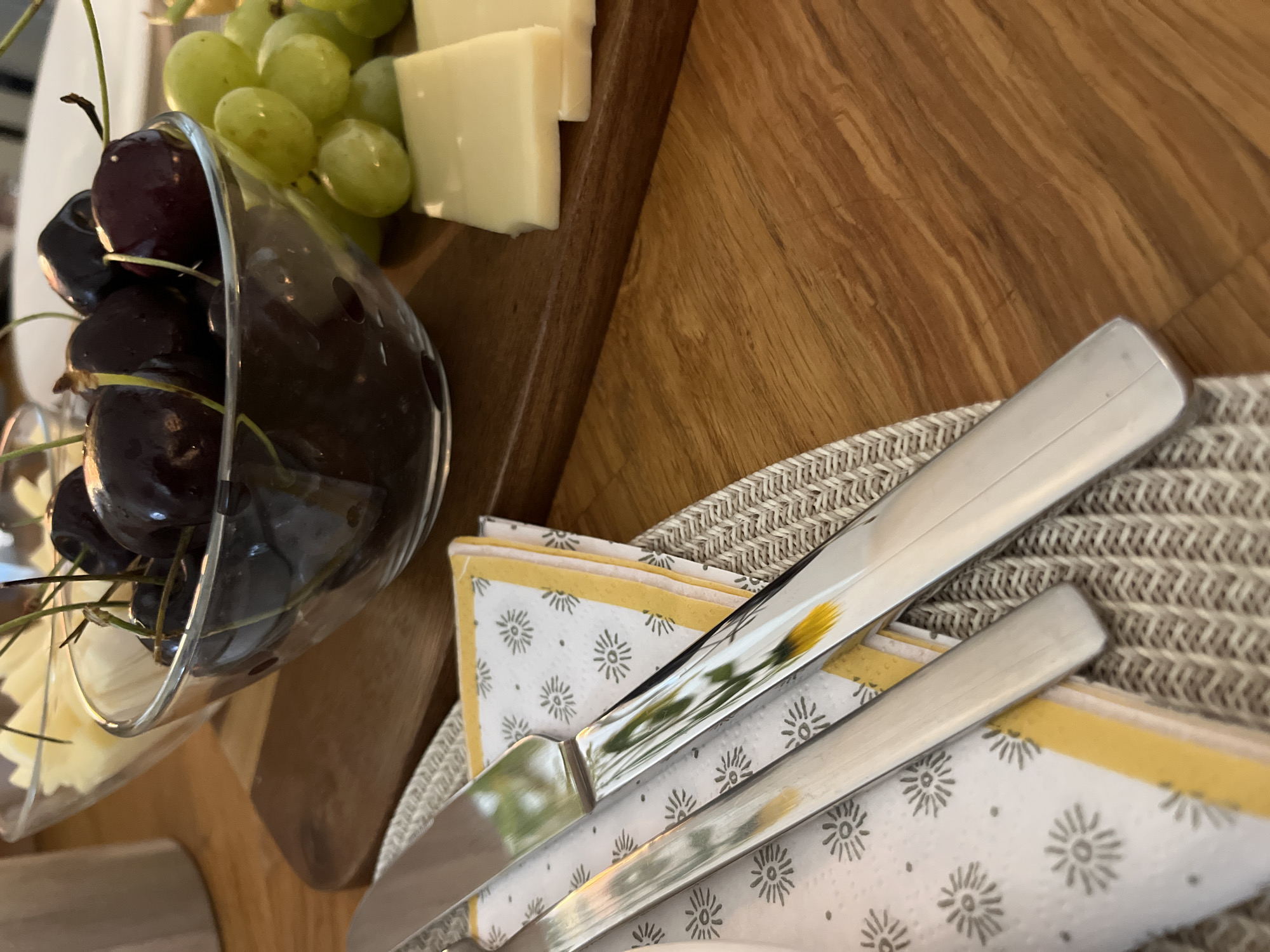 A close-up, high-angle shot shows a table setting with a variety of food items, including dark red cherries in a glass bowl, green grapes, and slices of yellow cheese. A wooden cutting board is partially visible beneath the food. To the left, a patterned napkin is placed on a woven placemat, with two polished metal utensils resting on top of the napkin. The background is blurred but suggests a wooden surface, possibly a table. The scene is set indoors, with soft, diffused lighting. There are no people visible in the image. The location is not specified in the image or implied by any of its contents.