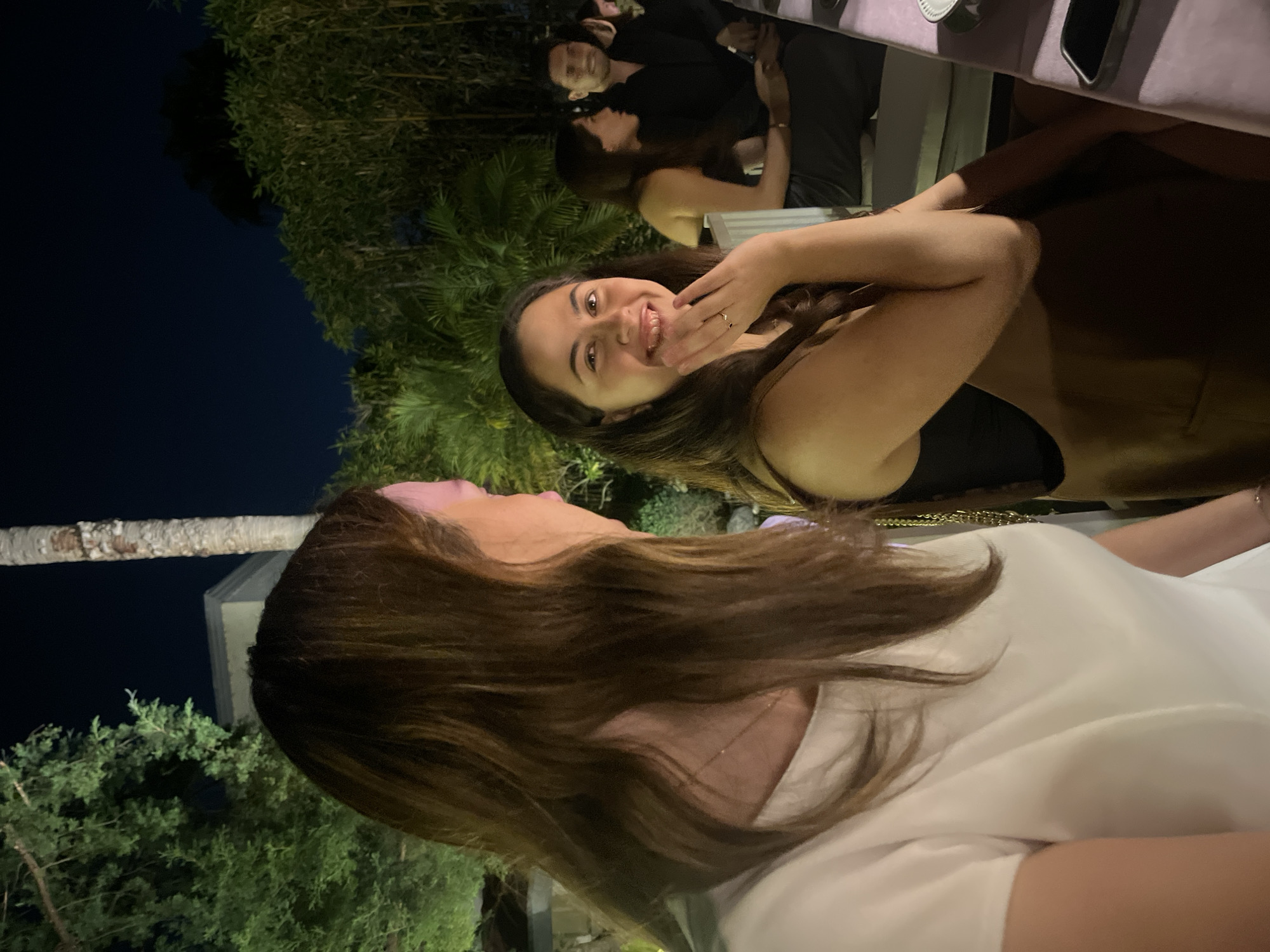 The image shows two women seated at a table outdoors at night. One woman is wearing a white shirt and the other woman is wearing a black top and gold dress. The woman in the black top is smiling and holding her hand up near her face. Behind them, there are other people at a table, as well as plants and trees. The location is San Bernardino Tlaxcalancingo, Mexico.