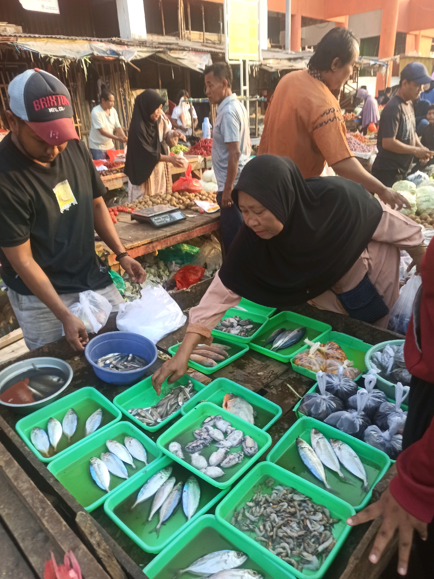 This is a medium shot, eye-level, outdoor photograph taken at a fish market, possibly in Cikarang, Indonesia. The image depicts a busy marketplace scene with several people and a variety of fresh fish displayed for sale.
In the foreground, a person wearing a black t-shirt and a "Brixton" cap is standing behind a table laden with green plastic trays filled with fish. Their hands are visible, interacting with plastic bags. To the right, a woman wearing a black hijab and a beige top is actively engaged in sorting or selling fish from a green tray. Her right hand is reaching into a tray of small fish.
Behind the foreground individuals, other people are visible, including a man in an orange shirt, a person in a light blue shirt, and another in a dark blue cap. They appear to be customers or vendors. The background shows stalls and structures typical of an outdoor market, with some wares, including red chilies and other produce, visible on tables. The ground is wooden and appears wet in places.
The fish are arranged in numerous green plastic trays. Some trays contain whole fish of varying sizes and types, while others hold smaller fish or seafood. There are also several plastic bags filled with what appears to be fish or other seafood. A blue bowl filled with small fish is placed on the table. A digital scale is also visible on the table in the middle ground. The lighting suggests it is daytime, possibly morning or late afternoon, given the warm tone of the light.