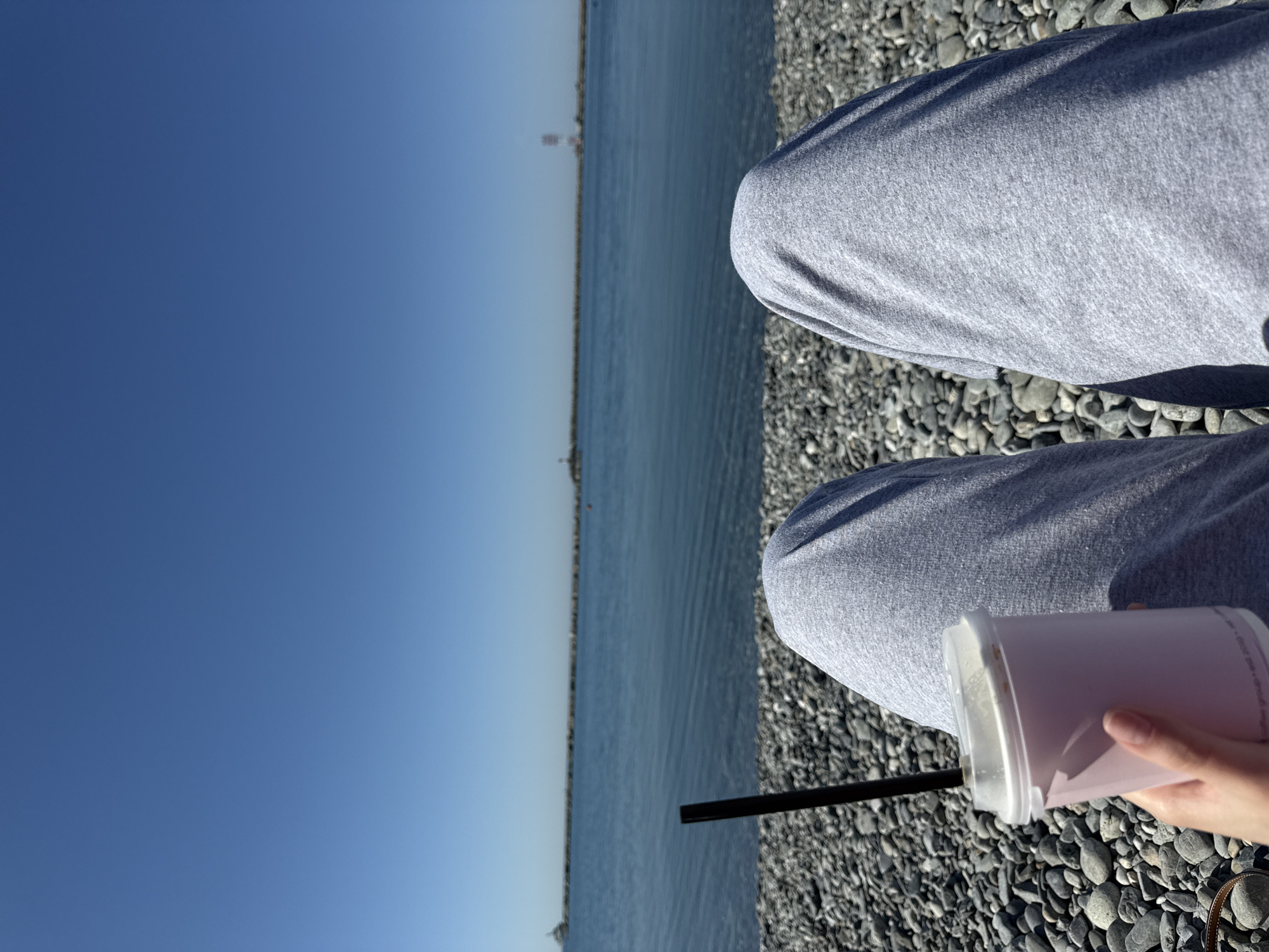 The image displays a first-person perspective, looking out towards a vast body of water on a sunny day. In the immediate foreground, the lower legs of a person are visible, clad in grey sweatpants. The individual is seated on a ground covered with numerous smooth, dark grey pebbles. A hand, likely belonging to the person, holds a tall, light pink or purple beverage in a clear plastic cup, which has a white lid and a black straw, resting on one of the legs.
The mid-ground features a wide expanse of calm, blue water, likely a sea or a large lake, stretching horizontally across the frame with gentle ripples on its surface. On the distant horizon, several indistinct structures, possibly buildings or towers, can be faintly seen. The upper portion of the image is dominated by a clear, bright blue sky, indicating fair weather. Shadows are visible on the person's legs and on the pebbled ground, suggesting strong, direct sunlight. Faint text, including the number "2019", is discernible on the side of the plastic beverage cup. The overall scene conveys a sense of relaxation by the water.
