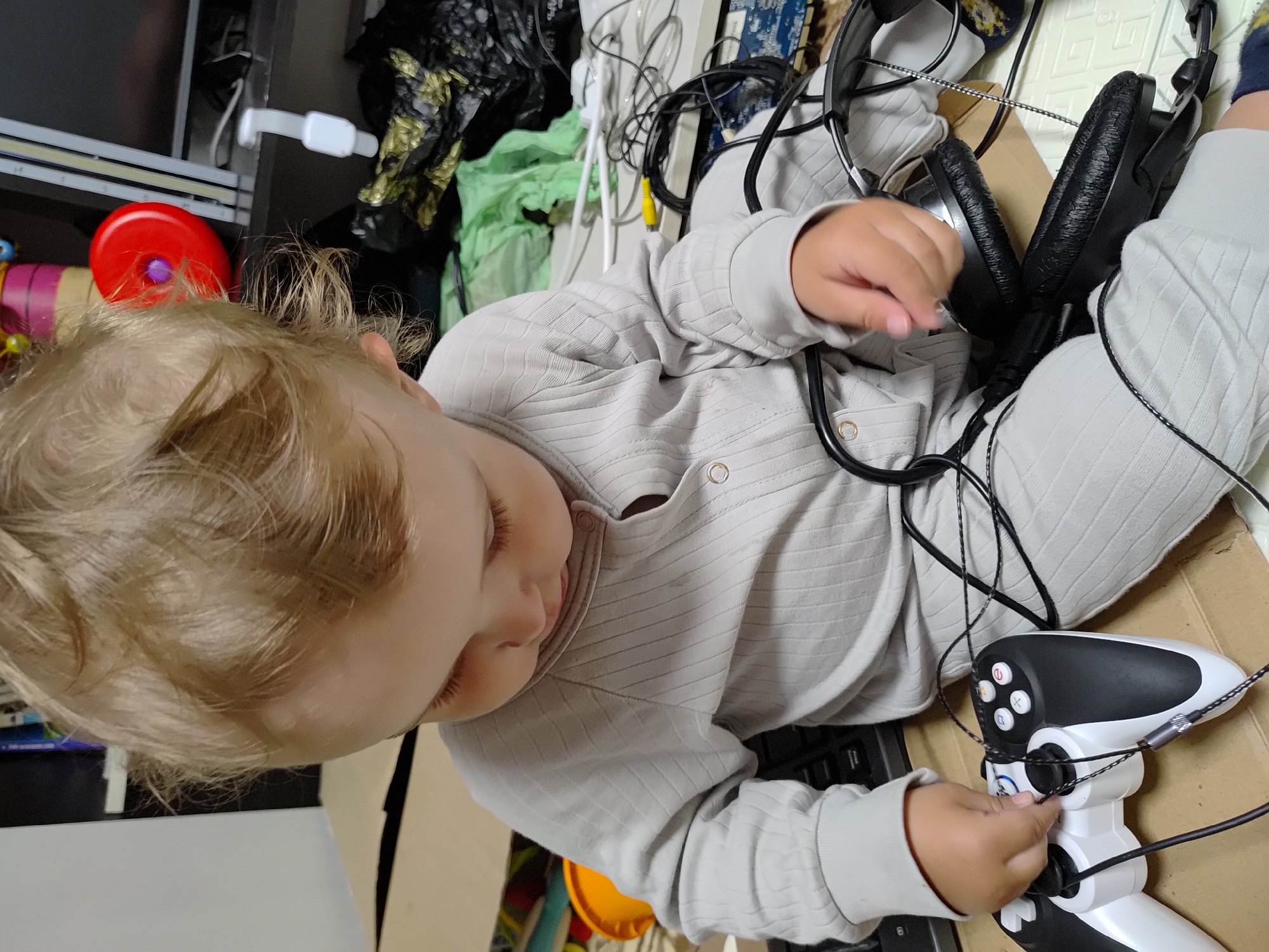 A toddler with blond hair is lying on their back, surrounded by various objects including headphones, a game controller, a keyboard, and numerous cables. The child is wearing a light-colored, long-sleeved shirt and pants. The background includes clutter and what appears to be a computer monitor. The location is identified as Марковский, Russia, though this cannot be independently confirmed from the image.