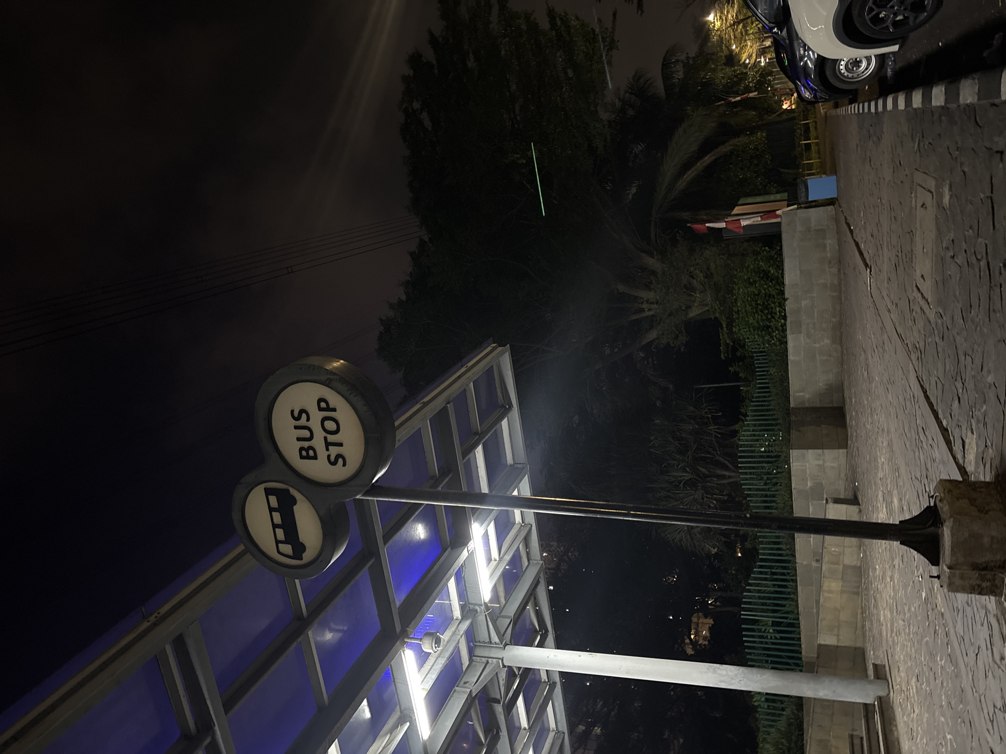 An outdoor, eye-level, night-time shot shows a bus stop sign with a bus silhouette and the words "BUS STOP" on it. The sign is mounted on a pole. To the left, a covered structure with blue lighting and white lights is visible, with a security camera mounted on the ceiling. The ground is paved with cobblestones. In the background, there are trees, a green fence, and a wall. To the right, cars are parked, and a trash can is present. The scene is likely located in North Jakarta, Indonesia, given the presence of the Indonesian flag.