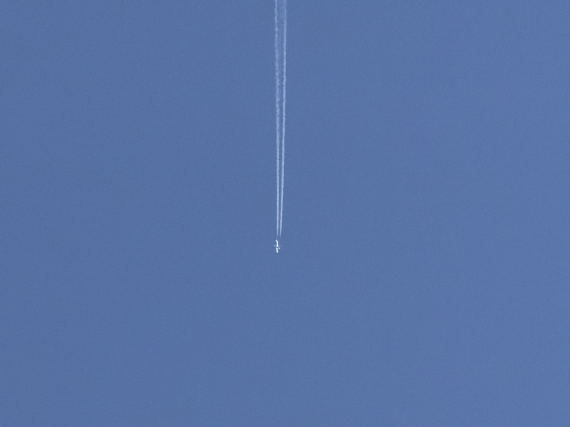 A distant commercial airplane is captured against a clear, blue sky, leaving a white contrail. The airplane is positioned in the center of the image.
