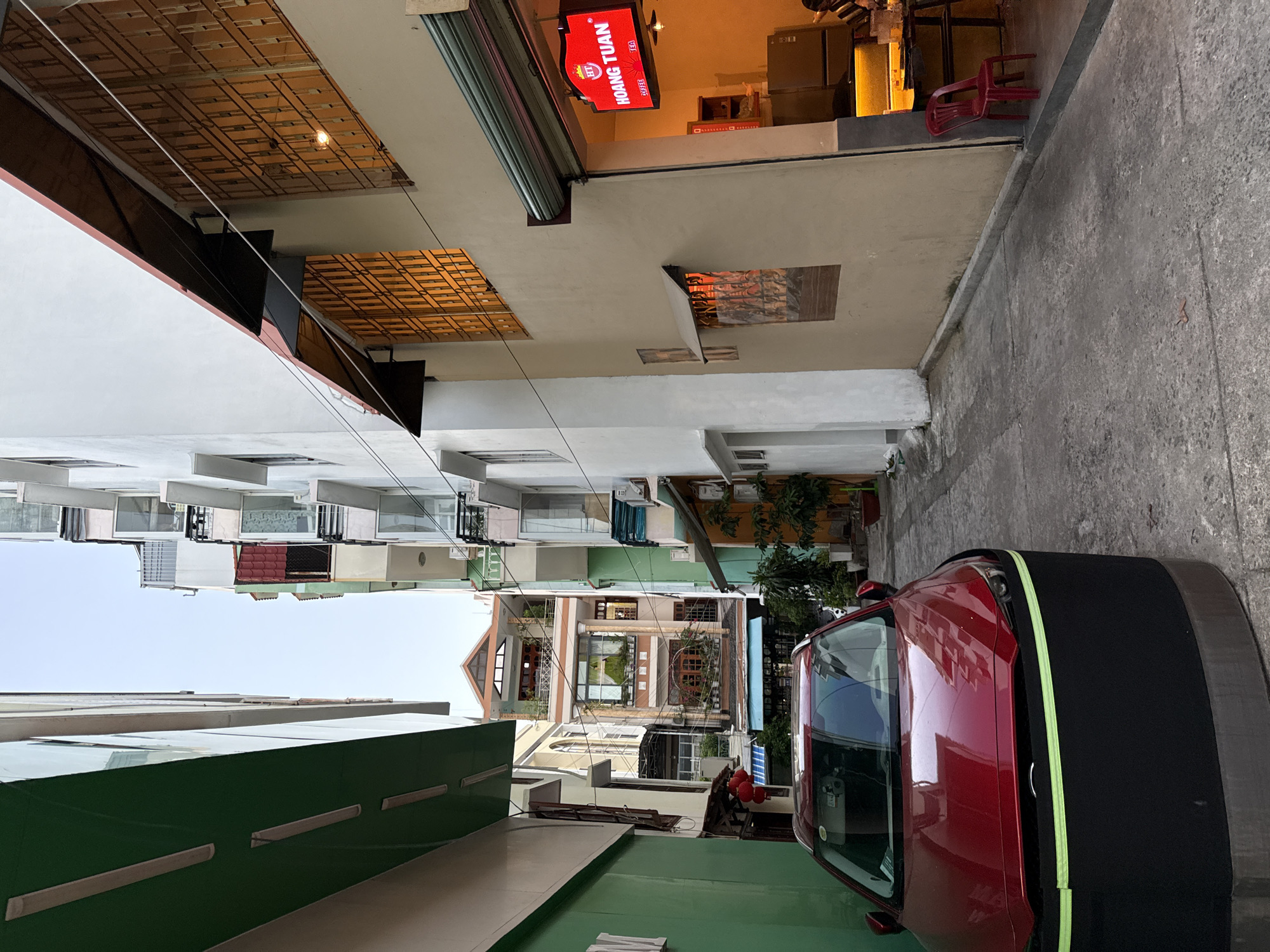 The image depicts a narrow alleyway in Nha Trang, Vietnam, flanked by buildings on both sides. A red car is parked in the foreground, partially obscuring the alleyway. On the right side of the alley, a building features an open storefront with a red neon sign that reads "HOANG TUAN COFFEE TEA." Inside, a counter and shelving are visible, illuminated by warm lighting. A red plastic chair sits outside the storefront on the pavement. The buildings in the alley are multi-story, with balconies, windows, and air conditioning units. One building on the left is painted green, while the building on the right is a light tan color. The sky is visible above, appearing clear and pale. The ground is paved with concrete, and a few potted plants are visible near the buildings.