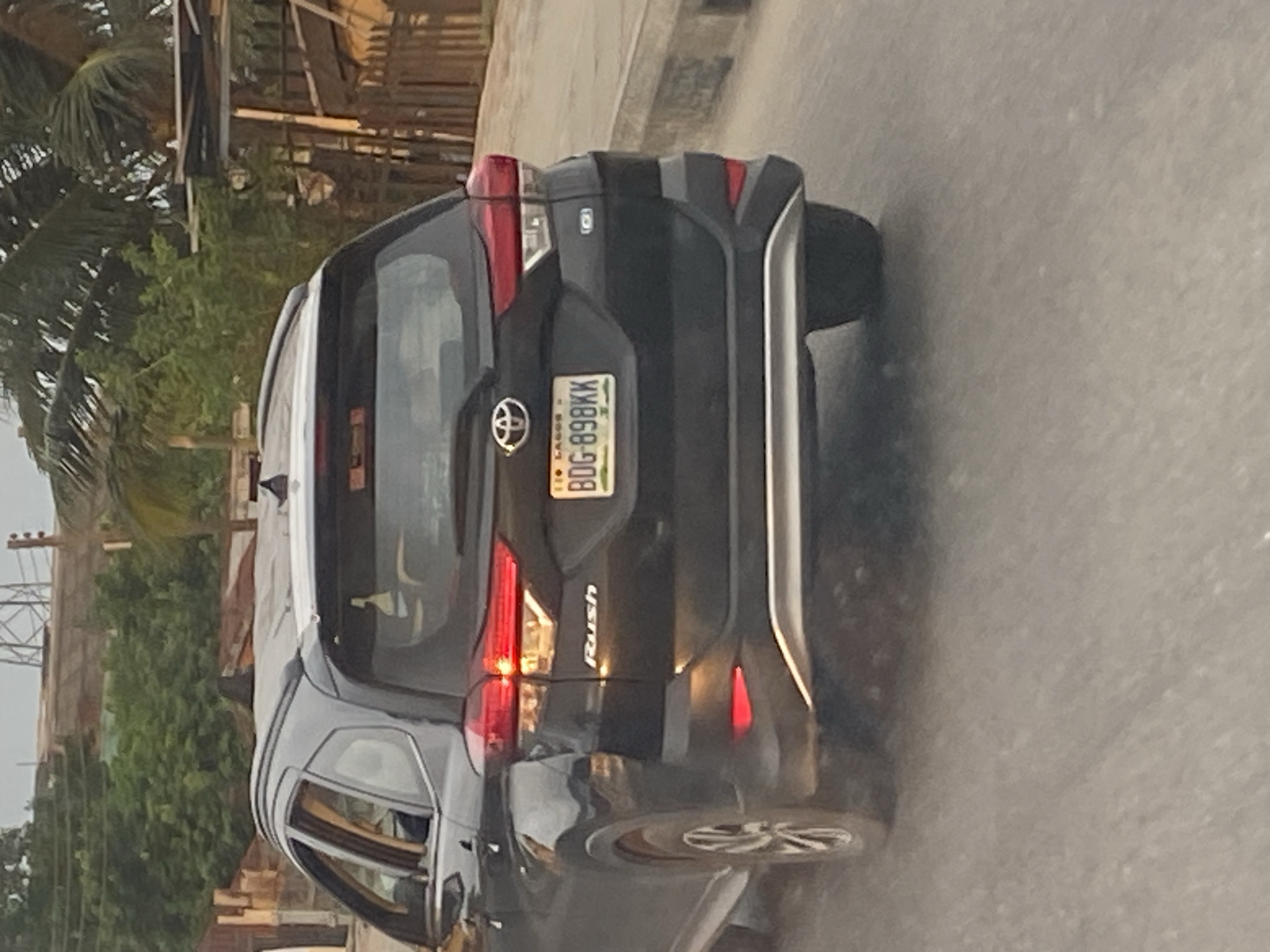 The image shows the rear view of a black Toyota Rush SUV in motion. The car is driving on a paved road in Ikeja, Nigeria. The license plate is visible and reads "BDG 898KK". The car has a silver trim on the rear bumper and red reflectors. In the background, there are trees, buildings, and a power pylon, indicating an urban environment. The lighting suggests it is daytime, possibly late afternoon due to the warm tones. No people are clearly visible inside or outside the vehicle. The scene is a typical street view within the city.