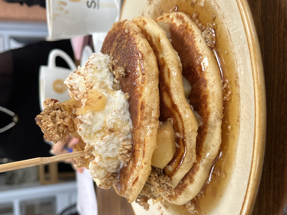 The image displays a close-up, eye-level view of a stacked pancake dish served on a round, light beige plate with a brown rim, resting on a dark brown wooden table. This scene is located within a cafe or restaurant in Skala, Greece.
The dish features three golden-brown pancakes, generously topped with a large, irregularly shaped dollop of white whipped cream. The cream and pancakes are dusted with a light brown powder, likely cinnamon. Visible on top of the cream and between the pancake layers are diced, yellowish-brown pieces of caramelized apple and clusters of light brown granola. A substantial amount of clear, viscous syrup or honey is drizzled over the entire stack, pooling at the base of the pancakes on the plate. A light brown, slender wooden skewer or pick is inserted vertically into the center of the topmost pancake.
In the blurred background, the upper torso of a person is visible, wearing dark clothing and a silver-colored chain or necklace. A hand, possibly holding a pen or utensil, is also discernible. Two white ceramic mugs are present; one to the left features a faint circular logo, while another is partially visible to the right.