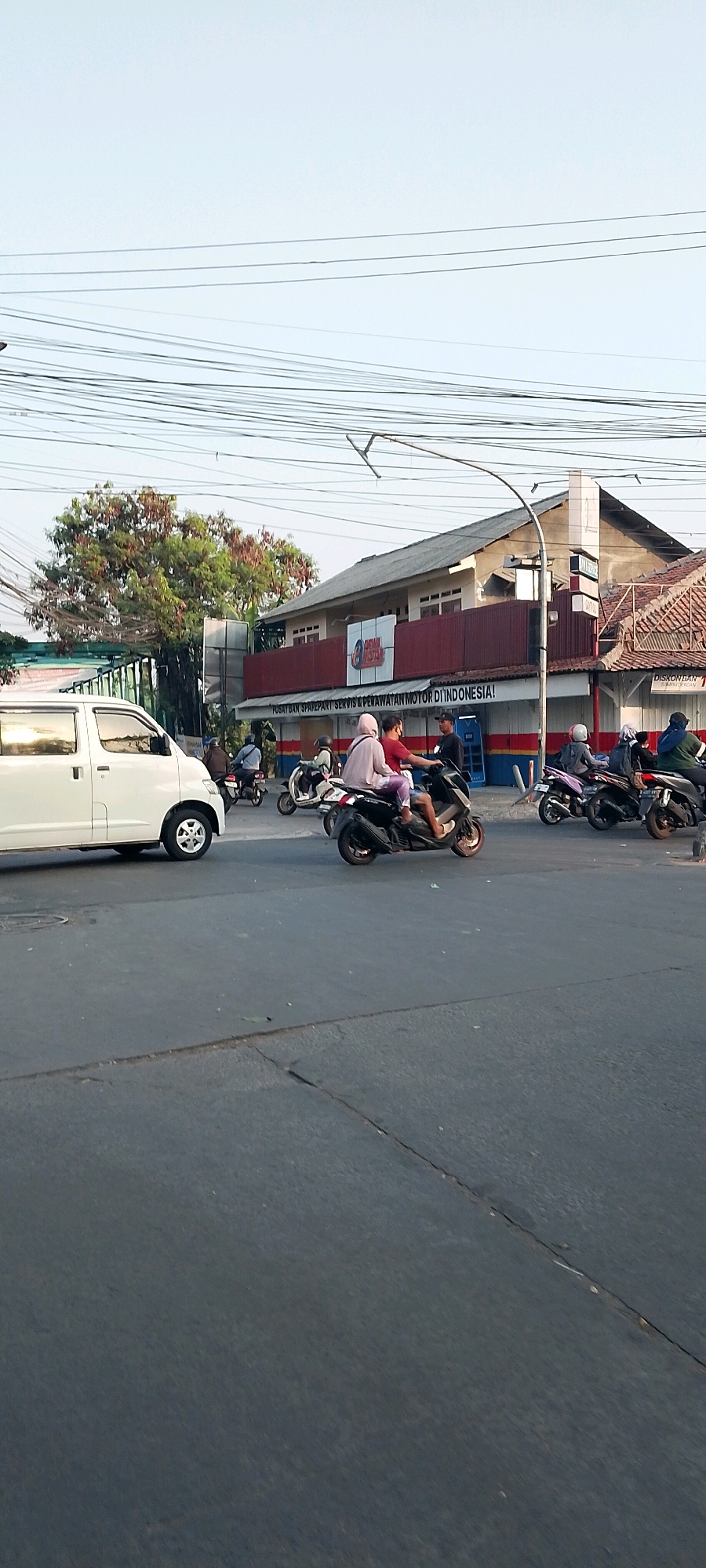 In Karangsatria, Indonesia, a street scene unfolds with a white van on the left side of the image. In the middle ground, several motorcycles are visible. Two individuals are riding on a black scooter, with one driving and the other seated behind. Another scooter is parked nearby, and further to the right, two more scooters are present, one carrying two passengers and another with a single rider. Behind the motorcycles, a building with a red and white facade is situated, featuring a sign that reads "PUSAT DAN SPAREPART SERVIS & PERAWATAN MOTOR DI INDONESIA!". Power lines crisscross the sky above the scene, and a tree with green and brown foliage stands to the left of the building. The road is paved and appears to be a junction, with a traffic light pole visible. The overall lighting suggests it is daytime, possibly late afternoon due to the warm tone of the light.