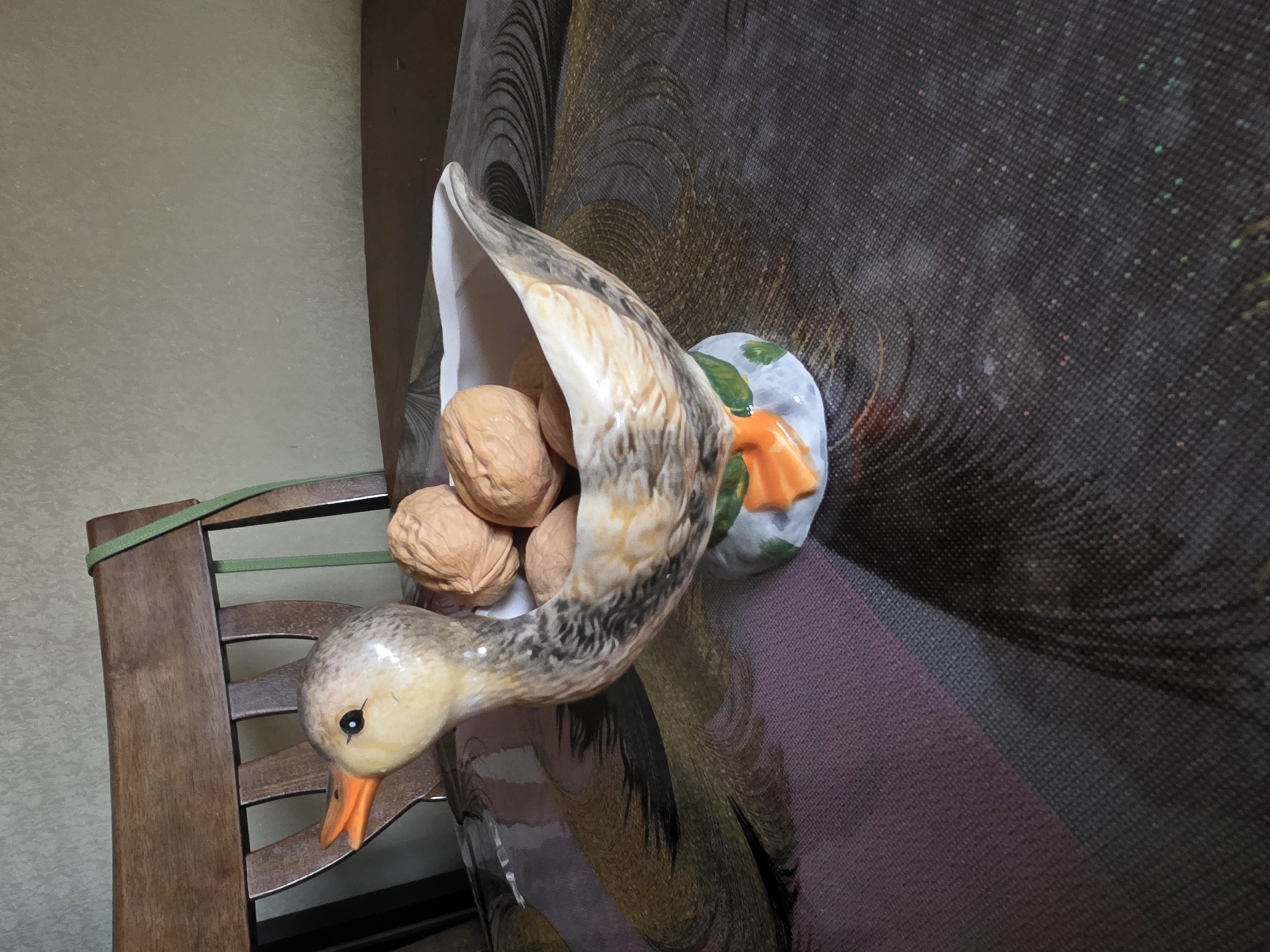 A ceramic duck figurine filled with walnuts sits on a table covered with a patterned tablecloth. The duck is positioned to the left of the frame, with its head turned to the left, beak open. The figurine has a base shaped like orange duck feet and green leaves. Behind the duck, a wooden chair with a green strap is visible. The background consists of a textured wall. The scene appears to be indoors. The image does not provide any information about the location of Levkovo, Russia.