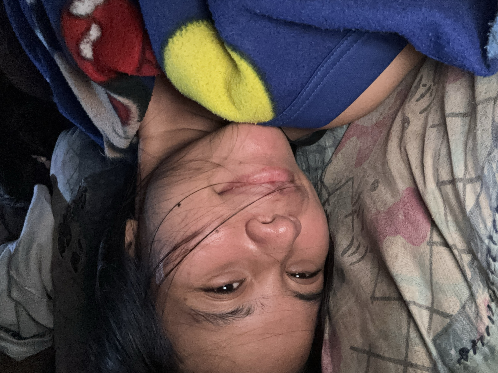 The image shows a close-up, eye-level, indoor shot of a young woman lying on her side in bed. She has dark hair and her eyes are open, looking towards the camera. Her face is partially obscured by a blue and yellow blanket, and the texture of the bedsheets is visible beneath her. The setting appears to be a bedroom. The image was taken in East Jakarta, Indonesia.