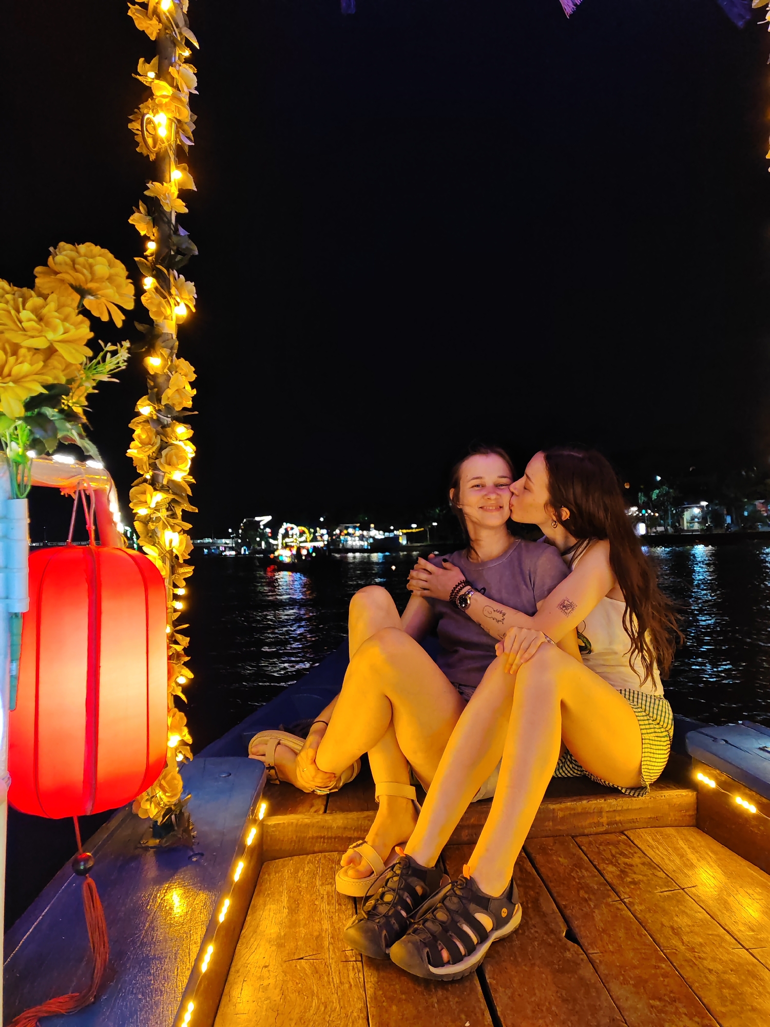 This image captures a tender moment between two women on a boat at night in Hoi An, Vietnam. The woman on the left, with short brown hair, is smiling as the woman on the right, with long dark hair, kisses her cheek. They are both seated on the wooden deck of the boat, their legs crossed and visible in the foreground.
The boat is decorated with a string of warm, yellow fairy lights intertwined with artificial yellow flowers, and a large red lantern hangs on the left side, adding to the festive atmosphere. The background shows a body of water with reflections of lights from buildings on the shore, suggesting a lively urban or tourist area at night. The darkness of the sky contrasts with the bright lights of the boat and the distant cityscape.
The overall mood is romantic and intimate, highlighted by the gentle kiss and the warm, inviting lights. The women are dressed casually, one in a grey t-shirt and the other in a white tank top and plaid shorts. The presence of tattoos on both women's arms adds a personal touch. The time of day is clearly night, and the atmosphere is festive and possibly celebratory. The water is calm, indicating mild weather conditions. No visible text is present in the image.