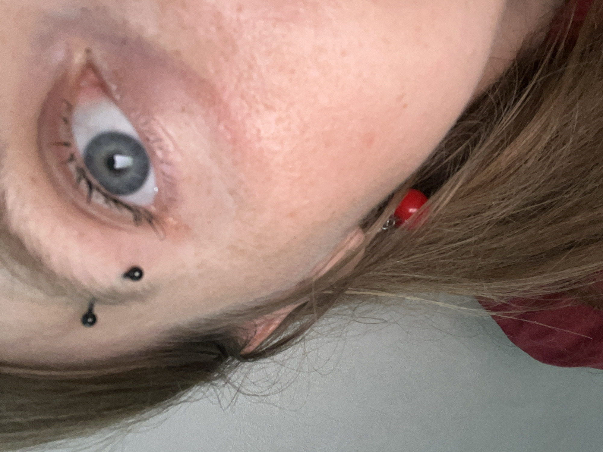 This is a close-up, low-angle, outdoor, daylight photograph of a young Caucasian woman's face, focusing on her eye and ear. She has fair skin with freckles, and light blue eyes. She is wearing two black eyebrow piercings and a red teardrop earring. Her hair is long and brown, and she is wearing a red shirt. The background is a plain, light-colored wall. The specific location within Сеніцкі сельскі Савет, Belarus, is not discernible from the image.