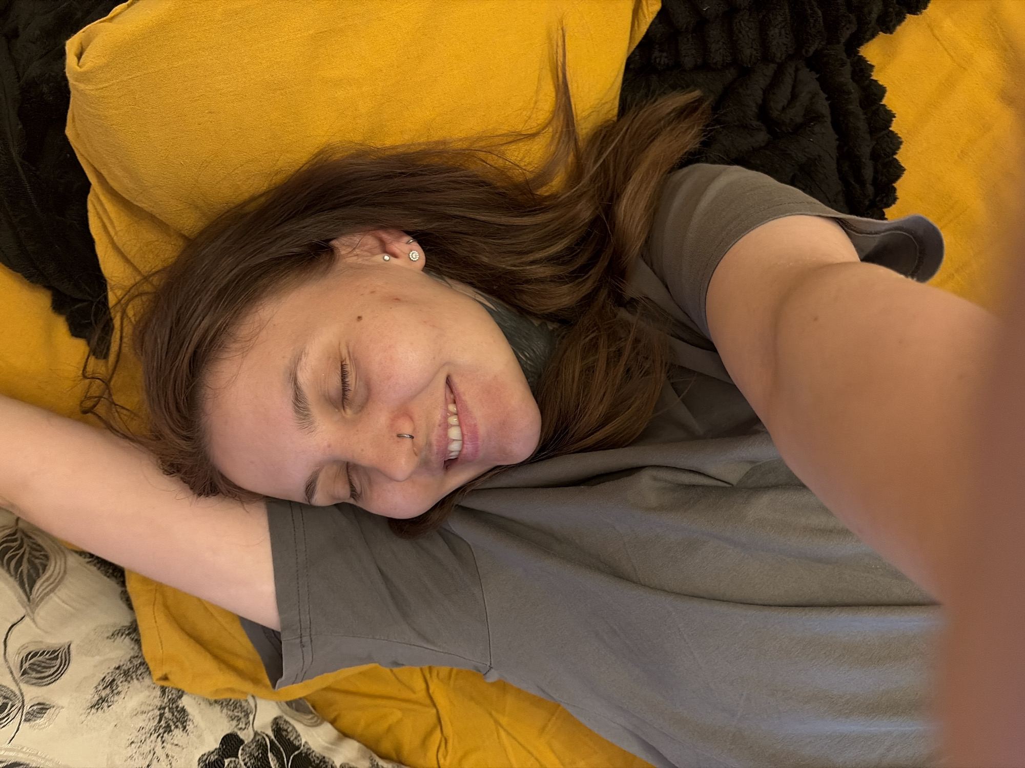 A woman is lying on her back on a bed. She has her eyes closed and is smiling. She is wearing a grey t-shirt. She has a nose ring and earrings. The bed is covered in yellow and black bedding. The background is out of focus. There is no information about the location in Chaikovsky, Russia.