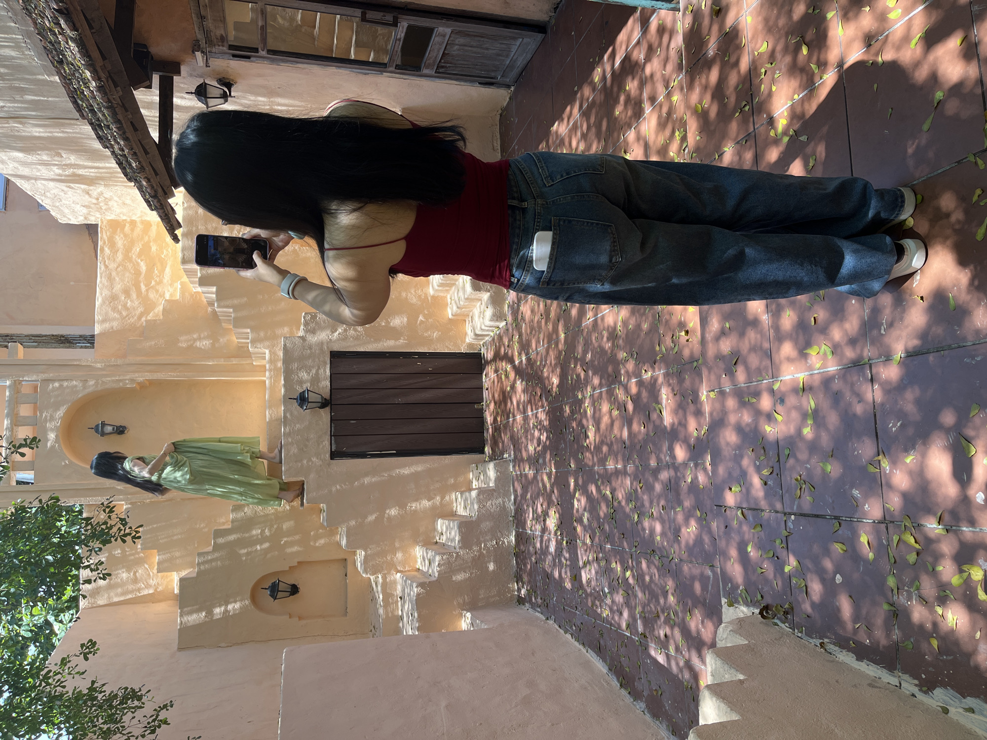 A woman is taking a picture of another woman in Đà Nẵng, Vietnam. The photographer is wearing a red tank top, blue jeans, and white shoes. The subject is wearing a long green dress. The women are standing on a beige-colored staircase. There are some leaves and tree shadows on the tile ground.