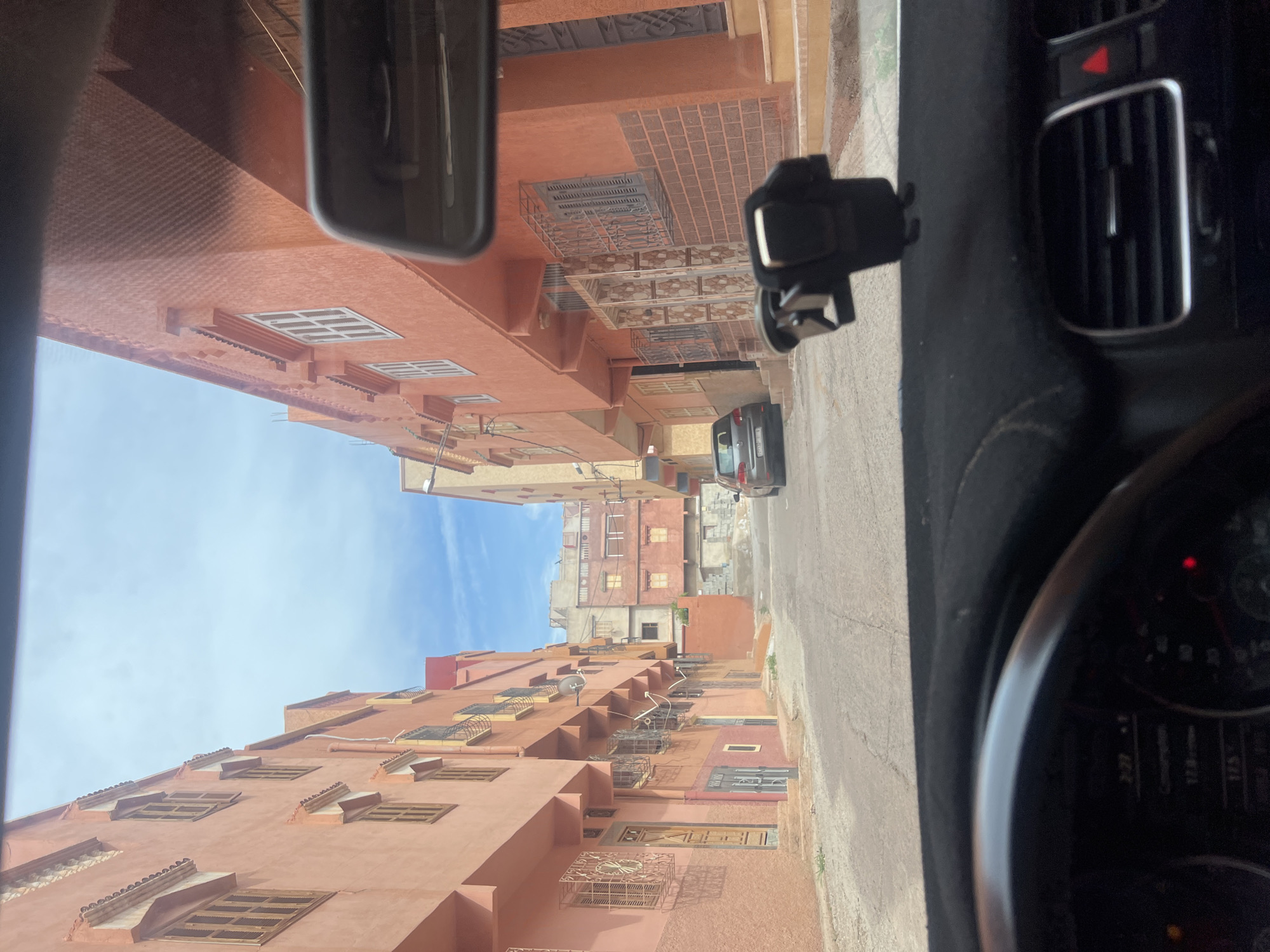 Captured from the interior of a vehicle, the image depicts a street scene in Ouaouizert, Morocco. The street is lined with buildings of similar architectural style and color. The perspective is from a vehicle, capturing the dashboard, steering wheel, and part of the rearview mirror. A car is partially visible on the street ahead. The sky is visible between the buildings, indicating a bright, daytime setting.