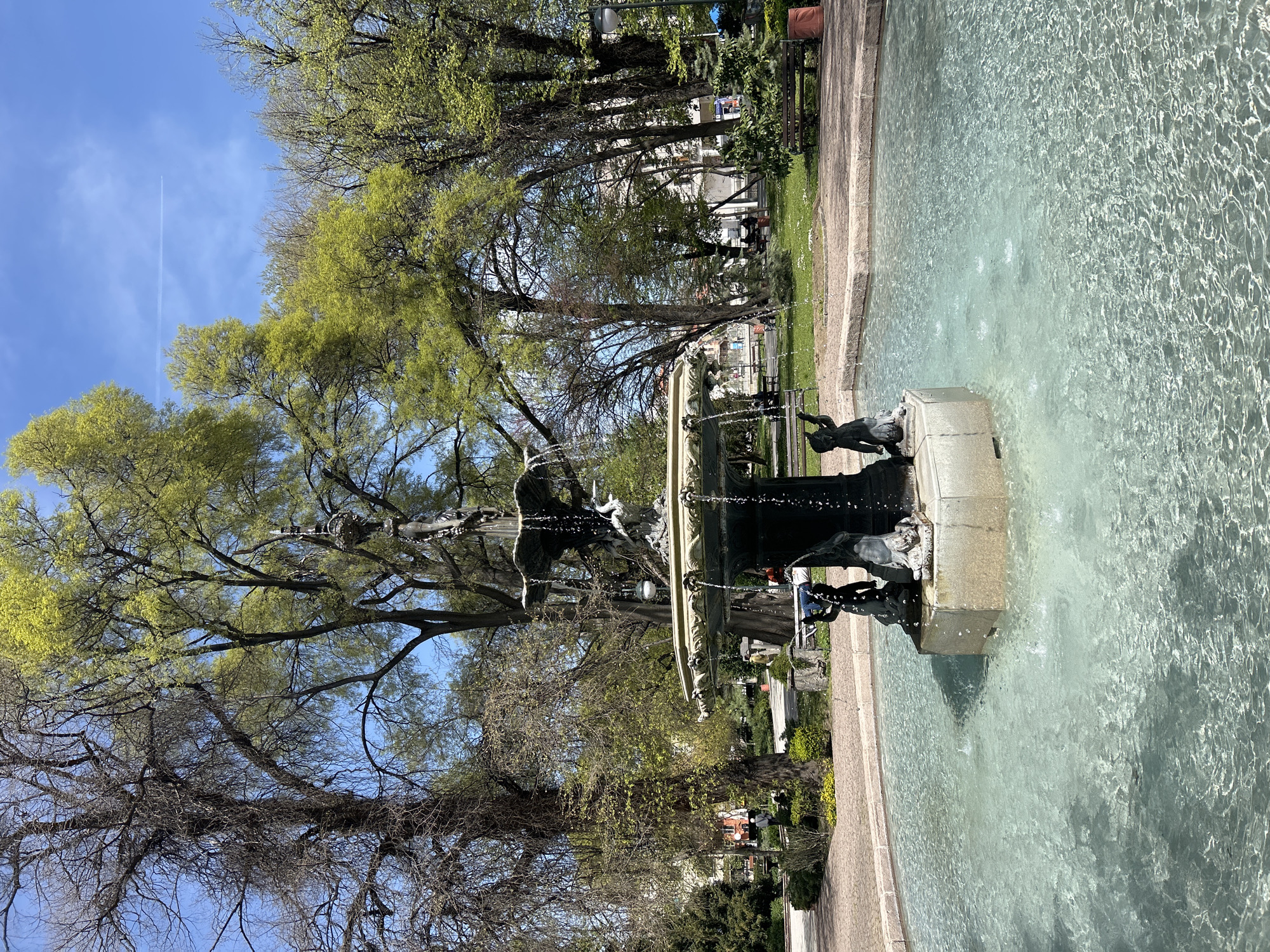 This vertical-format image depicts a serene public park scene in Plovdiv, Bulgaria, bathed in bright sunshine, likely during early to mid-spring. The central focus is a large, ornate fountain featuring multiple tiers and dark sculptural figures, possibly bronze, which appear to be cherubic or allegorical, accompanied by animals or mythical creatures. Water actively cascades from the fountain's various levels into a wide, shallow pool, creating ripples and reflecting the clear blue sky and surrounding greenery.
The fountain is encircled by numerous trees, some showcasing vibrant, newly emerged green leaves, while others still exhibit intricate bare branches. High above, a prominent white contrail from an airplane streaks across the bright blue sky. Paved pathways and grassy areas delineate the edges of the water feature. In the background, through the trees, park benches and distant architectural elements are discernible. A few people can be seen in the distance, relaxing on benches or strolling through the park. The scene is well-lit by direct sunlight, indicating a clear, pleasant day. No visible text is identifiable within the image.