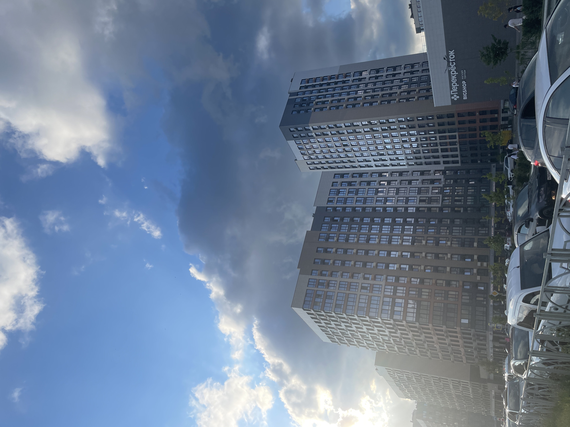 This image shows a street scene in Stavropol, Russia, featuring modern residential buildings. The buildings are tall and made of glass and concrete, with many windows. In the foreground, several cars are parked, and a metal fence is visible. The sky is partly cloudy, with dramatic dark clouds contrasting with patches of blue sky and bright sunlight, suggesting a dynamic weather condition or time of day, possibly late afternoon or early evening.
One of the buildings has a large sign that reads "Перекресток" (Perekrestok), which is a Russian supermarket chain. Below "Перекресток," smaller text indicates "BIGSHOP" and "МАГАЗИН" (magazin, meaning "shop").
There are a few people visible, mostly in the background near the buildings or cars, appearing to be pedestrians or shoppers. The overall atmosphere is urban and contemporary, with a focus on modern architecture and the daily life of a city. The lighting suggests a bright but potentially overcast day.