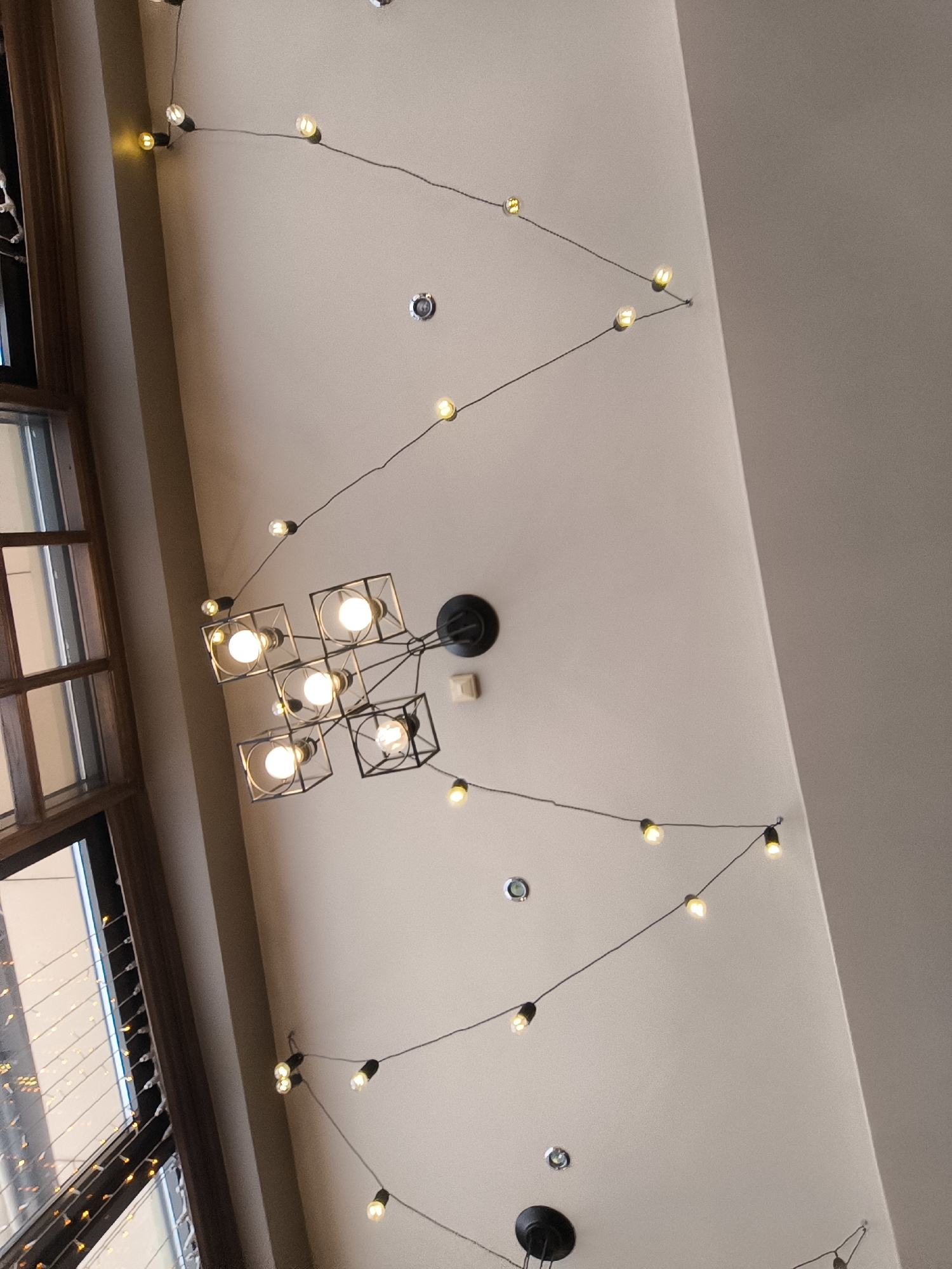 The image shows a ceiling with a geometric chandelier and string lights with small bulbs. The ceiling is a light beige color. In the upper left, there is a portion of a window with multiple panes. There are several ceiling fixtures visible in the image. The location appears to be indoors.