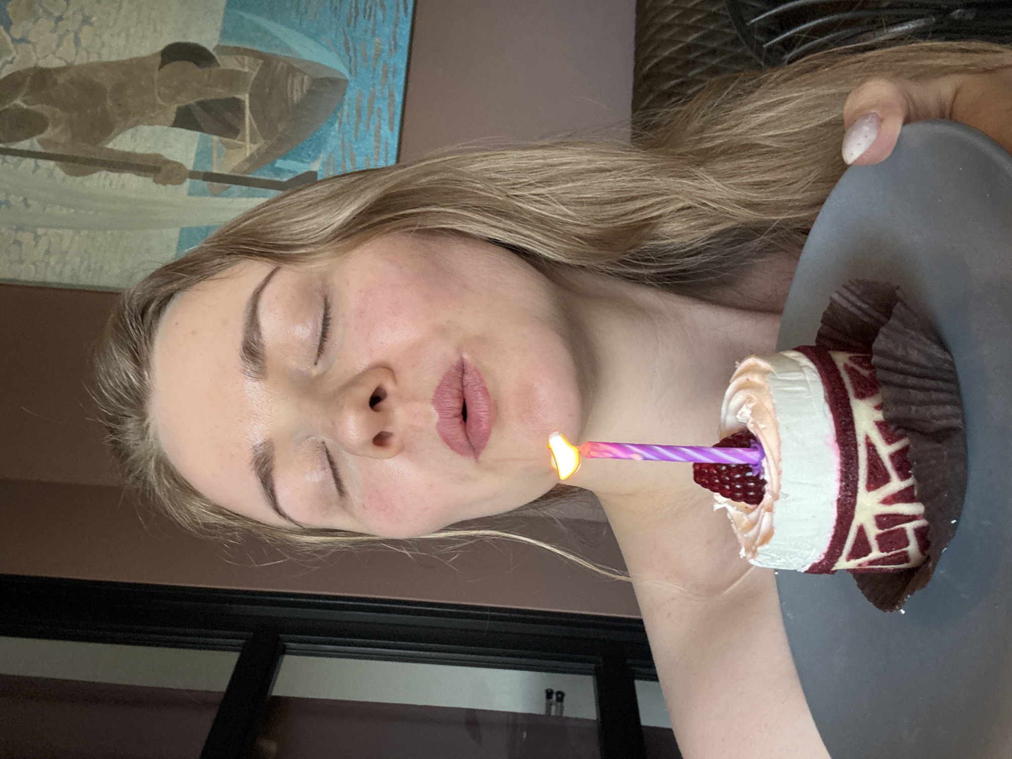 A fair-skinned woman with blonde hair is holding a gray plate with a cupcake. The cupcake has white and red frosting, raspberries, and a lit purple birthday candle. The woman has her eyes closed and appears to be blowing out the candle. A painting is visible in the background. The location is presumed to be indoors. There is no evidence to suggest that it is in the city of Saint Petersburg, Russia.