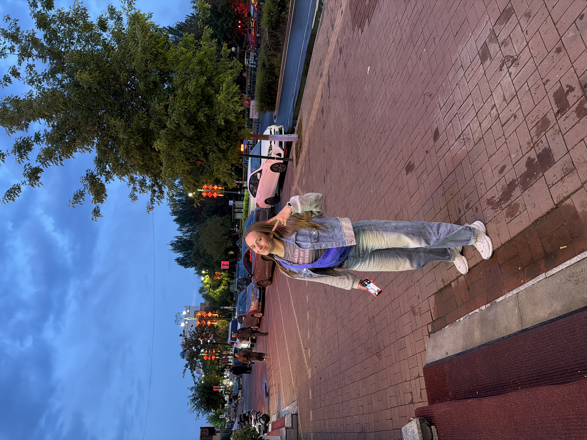 A young woman stands on a brick sidewalk in what appears to be a city street at dusk or early evening. The sky is a deep blue with visible clouds, and artificial lights from streetlights and buildings are on. She has long, light brown hair and is wearing a grey t-shirt, a blue denim jacket, light-wash jeans, and white sneakers, with a blue fanny pack across her chest. She is smiling directly at the camera and making a peace sign with her right hand, while holding a red smartphone in her left hand, which displays an image on its screen.
Behind her, a street is visible with several parked cars, including a white sedan. Beyond the street, there are numerous trees and buildings. Distinctive red, glowing lanterns hang from poles or structures along the street, suggesting an East Asian location, consistent with Tongjiang, China. Several streetlights illuminate the area, casting a warm glow. The sidewalk in the foreground is made of reddish-brown bricks, showing some darker, possibly wet, patches. In the lower right corner, a dark red textured mat is visible. The scene has a casual, urban feel with the woman as the central focus. Text visible on her t-shirt partially reads: "THE ART OF DRAMA PROCESS".