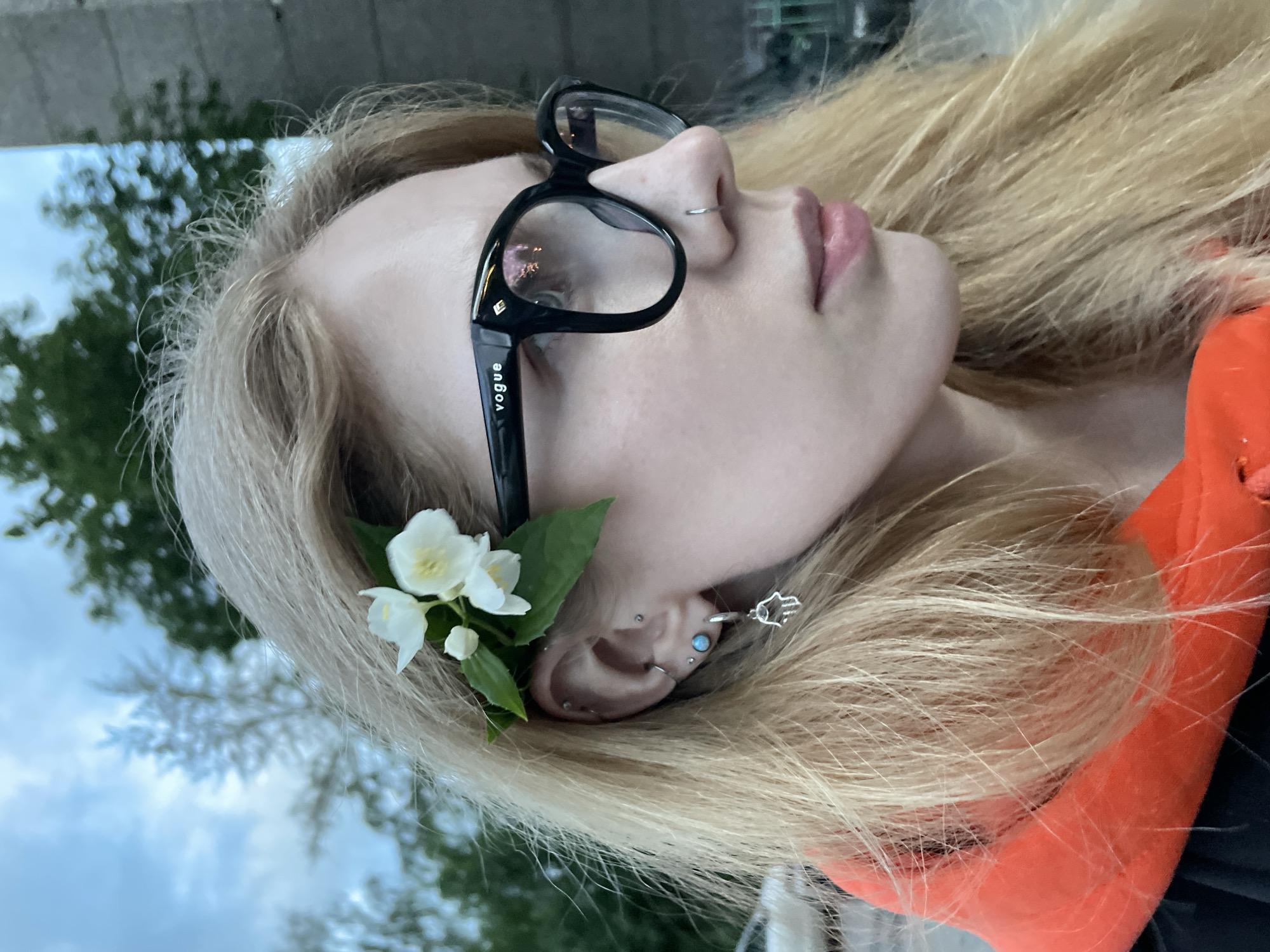 This close-up, sideways portrait features a young individual, likely female, with long blonde hair. She wears black-framed glasses, with the word "VOGUE" visible in white text on the right temple arm. A small silver hoop piercing is visible in her nostril. Her ear is adorned with multiple piercings, including an opal-colored stud and an earring shaped like a Hamsa hand. Tucked behind her ear is a cluster of small white flowers with green leaves, possibly jasmine. Her lips are slightly parted, and her eyes are either closed or cast downwards, giving her a serene or contemplative expression. A vibrant orange garment, possibly a hoodie, is visible on the right side of the frame. The background is softly blurred, showing an overcast sky with patches of green foliage, suggesting an outdoor setting in Izhevsk, Russia, during daylight hours.