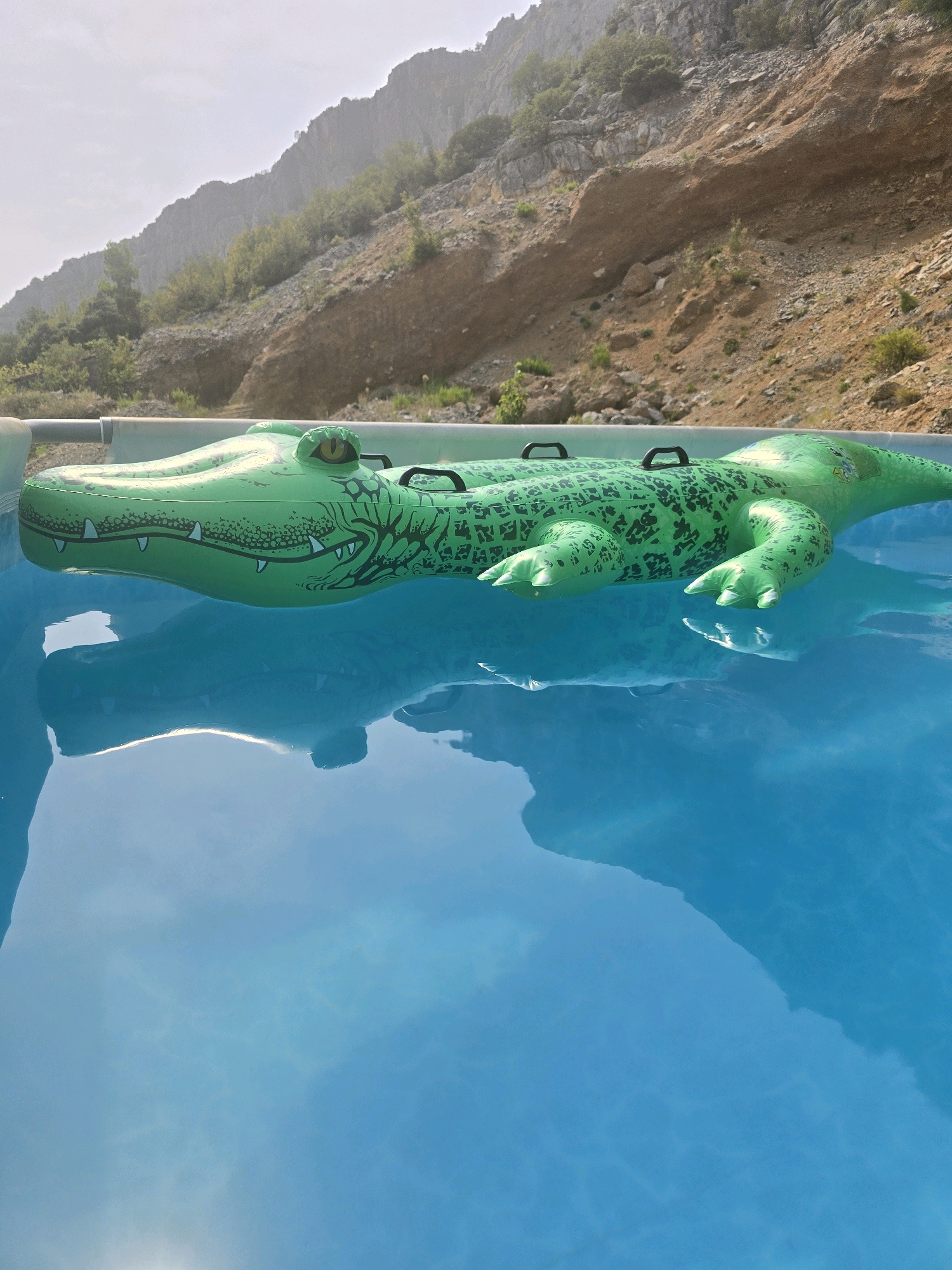 A green inflatable alligator pool float rests on the surface of a bright blue swimming pool. The pool is surrounded by a light-colored rim, and beyond it, a rocky, barren hillside rises under a hazy sky. The hillside is sparsely dotted with green vegetation. The scene appears to be in an outdoor setting, likely during the daytime. The specific location in the city of Podgorica, Montenegro, is not discernible from the visual information provided in the image.