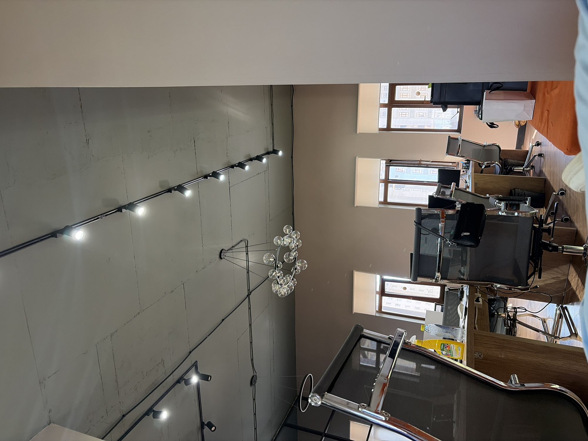 The image shows an office interior, likely in Astana, Kazakhstan, with a focus on the ceiling and upper parts of the room. Multiple track lighting fixtures are mounted on the exposed concrete ceiling, with several lights illuminated. A unique chandelier made of clear glass spheres is suspended from the ceiling. In the foreground, the metallic frame and mesh of an office chair are visible. The background reveals windows with blinds, and office desks with computers and chairs. The overall aesthetic suggests a modern, minimalist office design.