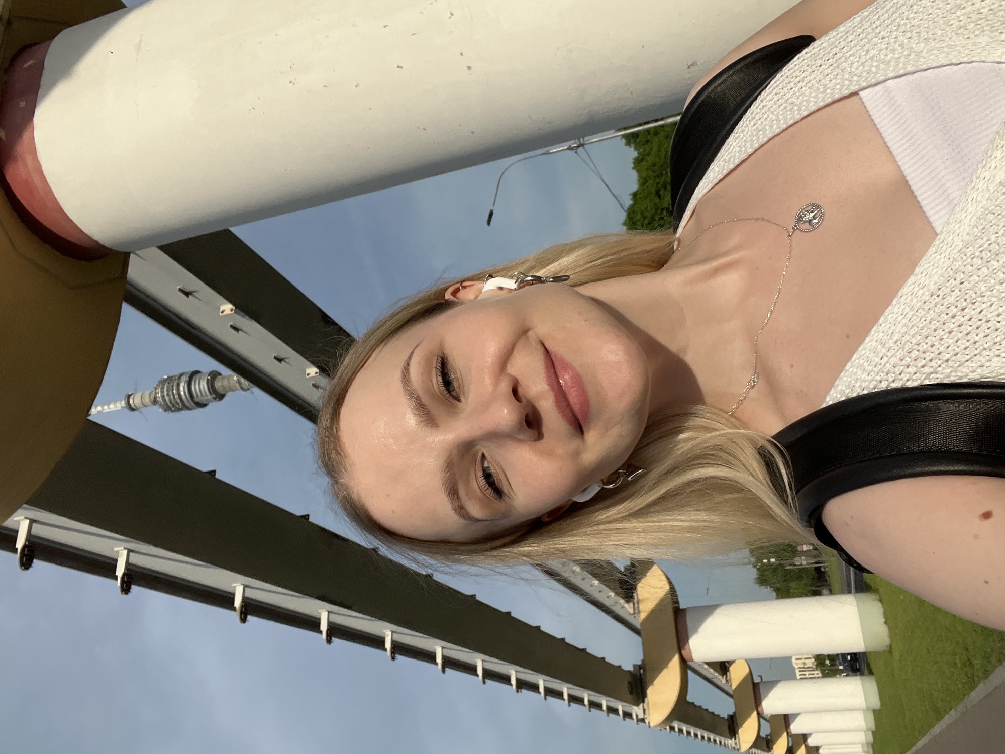 A fair-skinned woman with blond hair is centered in an overhead shot taken on a bridge in Moscow, Russia. The woman has light eyes, light eyebrows, and pink lips. She is wearing white wireless earbuds and a silver necklace that falls from her neck to the front of her white top and white cardigan. Her black purse strap is visible.
The bridge is painted with white pillars, some of which have a tan brace. Overhead, there are dark metal bars with silver hardware and white brackets. In the background, there is green grass, a street with traffic, and apartment buildings. The sky is a light, pale blue.