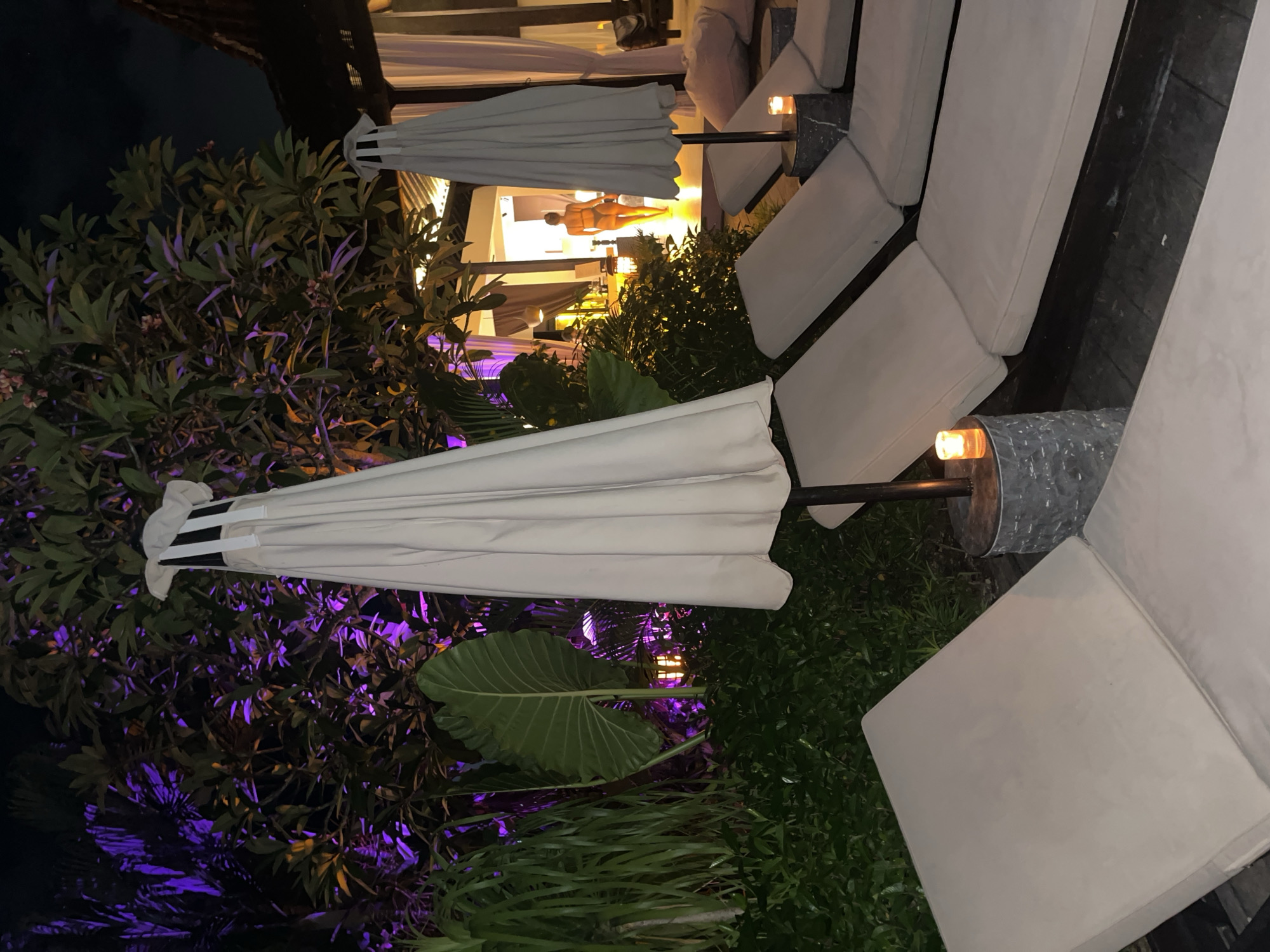 This image, taken at night in Mengwi, Indonesia, captures a luxurious outdoor lounge area. The scene features multiple comfortable white lounge chairs with matching cushions, arranged on dark wooden decking. Several large, closed white patio umbrellas are positioned among the seating and lush greenery. Small, glowing lights, likely candles or lanterns, sit on side tables next to some of the chairs, contributing to the ambient lighting.
Vibrant tropical foliage and bushes are prominent, with some sections dramatically illuminated by decorative purple and orange lights, creating striking color contrasts against the dark night sky. In the brightly lit background, through an open structure, a light-skinned mannequin or statue is visible, standing upright and facing away. It appears to be wearing a two-piece swimsuit or undergarment, its silhouette highlighted by the warm indoor lighting.
The overall setting suggests a tranquil and upscale resort or hotel environment, designed for evening relaxation. No specific activity or event is taking place, and no text is visible. The combination of comfortable furnishings, natural elements, and sophisticated lighting creates an inviting and peaceful ambiance.
