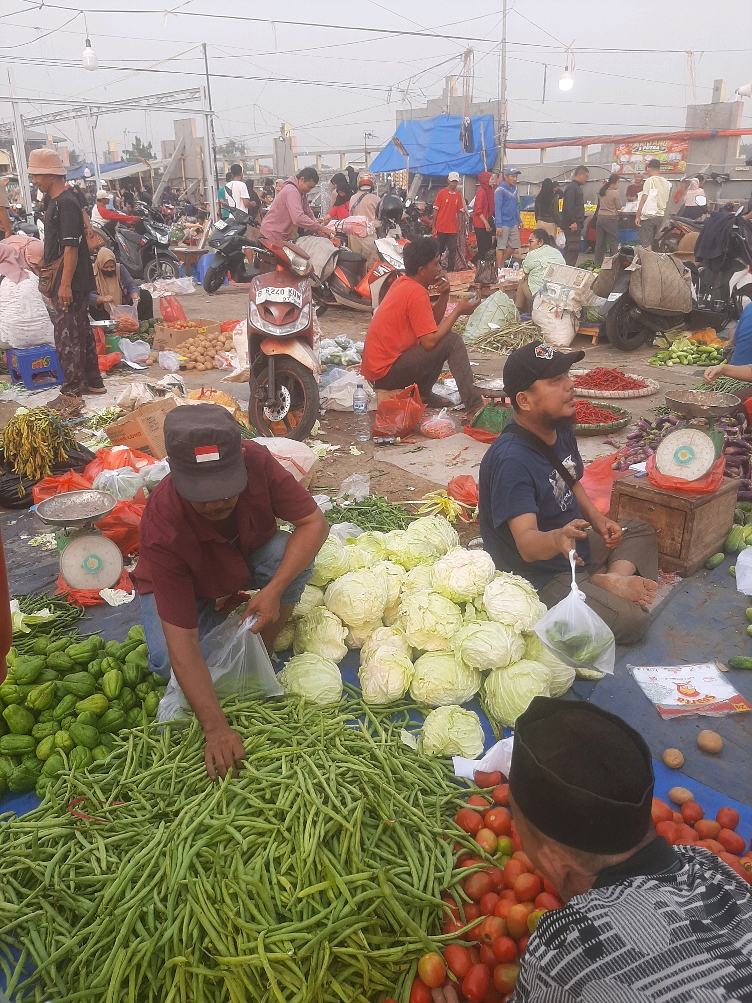 This image depicts an outdoor market scene, likely a traditional wet market, located in Bekasi, Indonesia. The market appears crowded and bustling, with numerous people engaged in various activities. In the foreground, a man in a maroon shirt and blue jeans is squatting, holding a plastic bag with vegetables, and surrounded by large piles of green beans, chives, and cabbages. To his right, another man wearing a black baseball cap and dark blue t-shirt is sitting on the ground, also holding a plastic bag of vegetables. In the background, there are more people, motorcycles, and stalls displaying a variety of produce, including tomatoes, potatoes, and chilies, as well as other goods. The environment is generally unkempt, with dirt and various items scattered on the ground. The overall impression is one of a vibrant, informal marketplace operating under a bright, overcast sky.