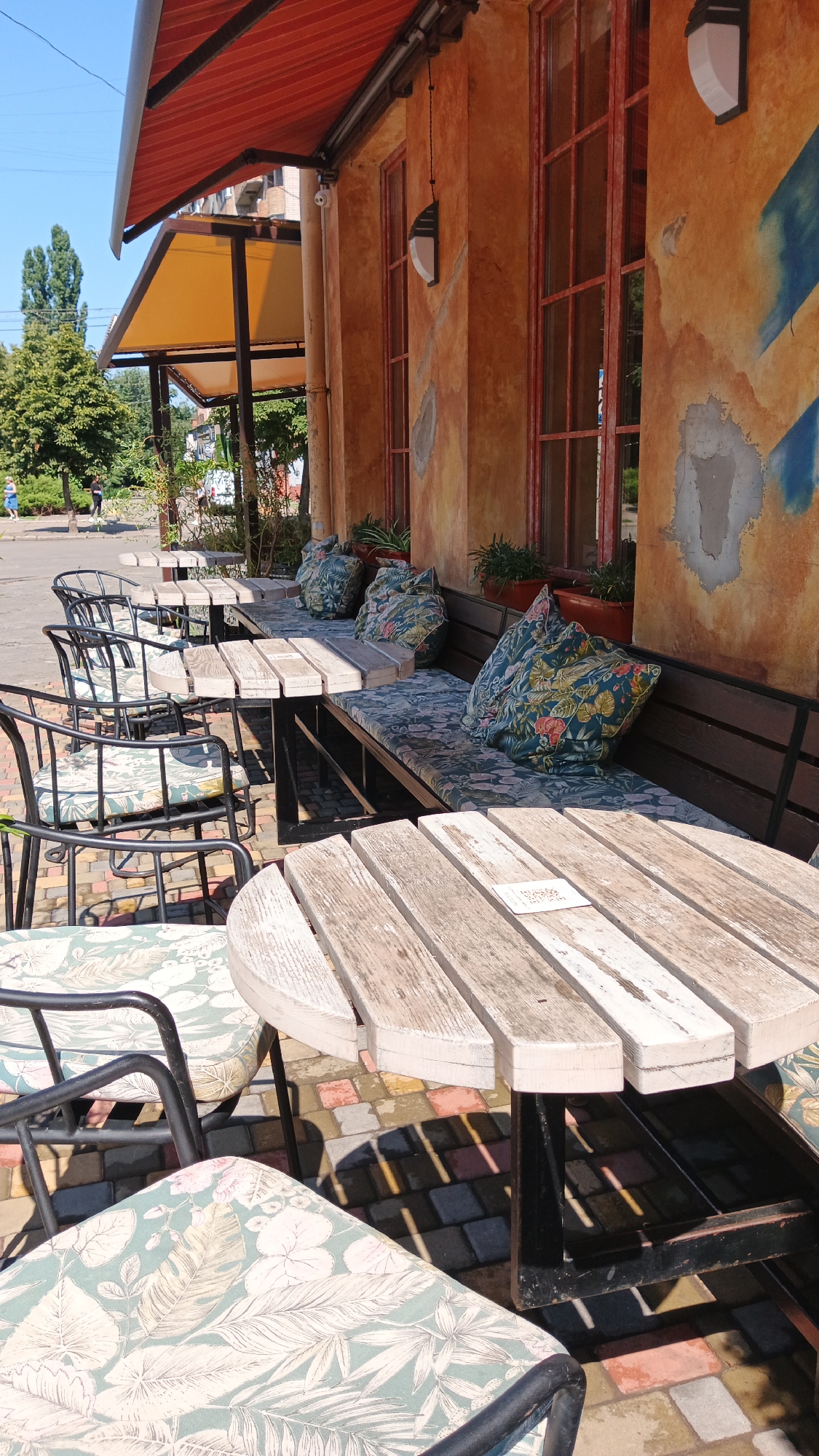 The image depicts an outdoor seating area for a cafe or restaurant, likely located in Kryvyi Rih, Ukraine, given the context. The scene is bathed in bright sunlight, casting strong shadows on the brick-patterned ground. In the foreground, a light-colored, weathered wooden table with a slatted top is positioned over a black metal base. Two chairs with decorative floral cushions, featuring a pattern of green leaves and white flowers, are visible next to the table.
Behind this, a dark wooden bench with matching floral cushions is situated against a building with a textured, ochre-colored wall. The wall has large, red-framed windows and shows signs of wear and abstract blue paint strokes. Several potted plants are placed along the windowsill above the bench. Further back, more black metal chairs with similar floral cushions surround another wooden table.
The background shows trees and a clear blue sky. A red awning extends over the seating area, and a yellow awning is visible beneath it. A few blurred figures can be seen in the distance, walking along a paved area adjacent to the cafe. The overall impression is of a pleasant, sunlit outdoor dining space.