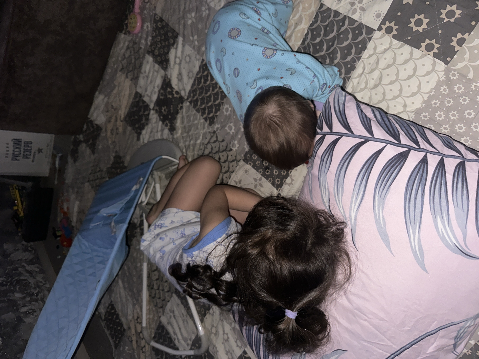 The image shows two children lying on a bed covered with a patterned quilt. One child is wearing blue pajamas with a repeating pattern and is partially covered by the quilt. The other child, closer to the camera, has dark hair pulled back into a ponytail with a purple hair tie and is partially covered by a pink blanket with a leaf-like pattern. A blue and gray folding bed is positioned next to them. A cardboard box is visible on the floor. The scene is indoors, likely in a bedroom. The location and city are not identifiable.
