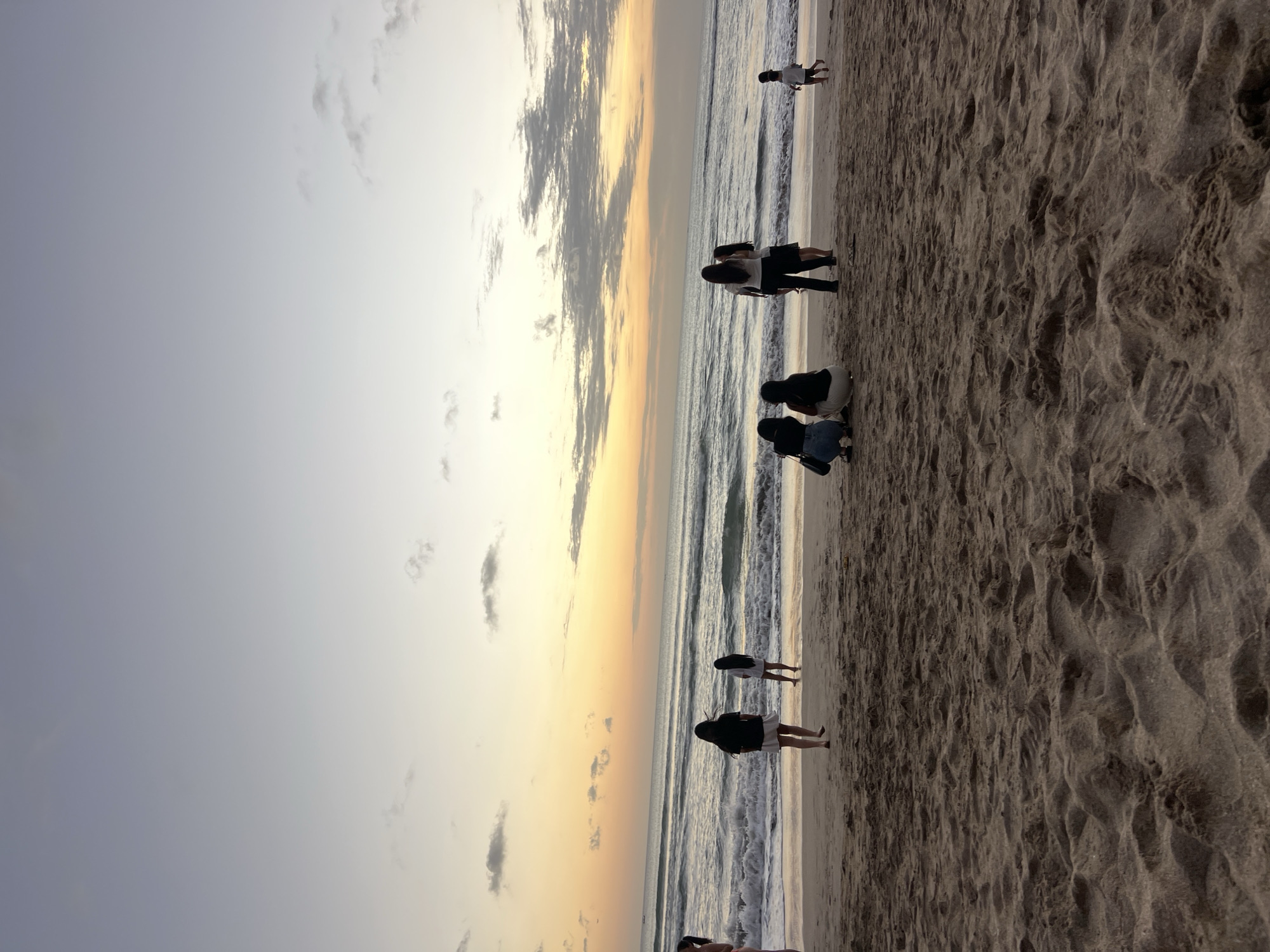 The image captures a scene on the beach in Kuta, Indonesia, during what appears to be sunset or dawn. The sky is a gradient of soft blues and yellows, with scattered clouds. Gentle waves are breaking on the shore, creating a foamy white line along the wet sand.
Several individuals are present on the beach. In the foreground, the textured sand is prominent, with footprints visible. Further back, near the water's edge, a group of people are gathered. Some are standing, while others are seated. One person is walking towards the water, and a child is seen running further down the beach.
The overall atmosphere is serene, with the soft lighting and the natural elements of the beach creating a peaceful ambiance. The individuals appear to be enjoying the scenery and the tranquil environment of Kuta beach.