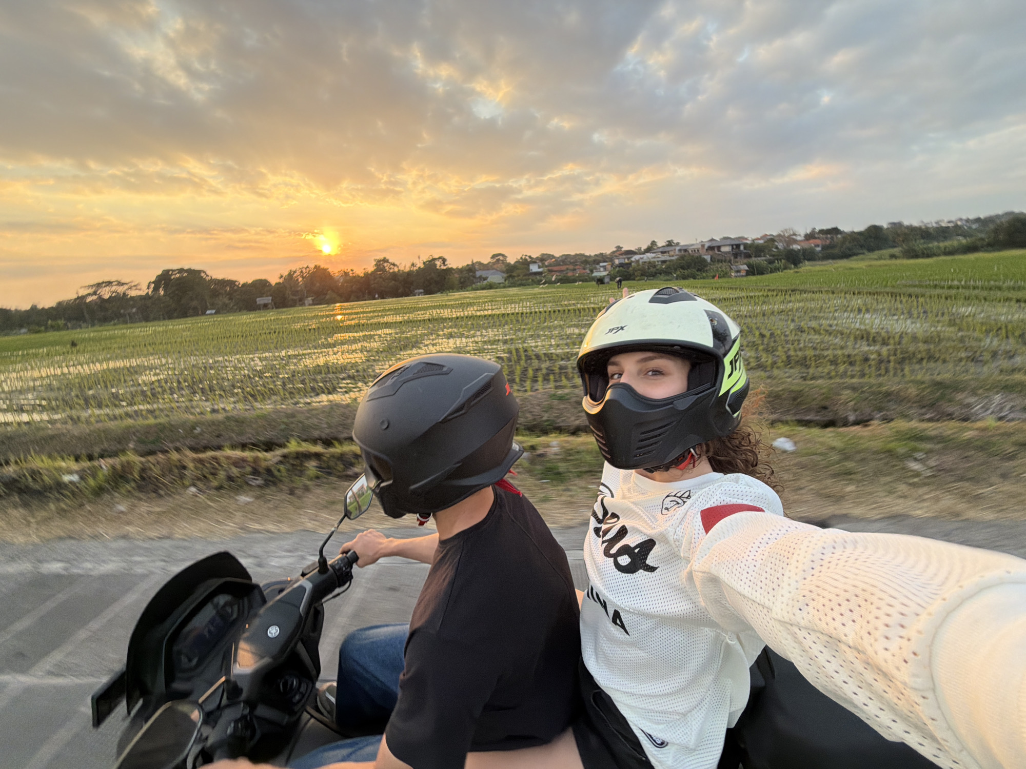 In a selfie taken from the perspective of a motorcycle rider in North Kuta, Indonesia, two individuals wearing full-face helmets are captured during what appears to be sunset. The rider in the foreground, facing the camera, wears a white helmet with green accents and a white long-sleeved shirt with red trim and black lettering. Their right arm extends towards the camera, holding the phone. Behind them, a second individual wearing a black helmet and a black t-shirt is visible, holding the motorcycle handlebars. The motorcycle itself, a dark scooter, is also partially visible. The backdrop features a blurred, vibrant landscape of rice paddies, with trees and houses lining the horizon under a dramatic sky filled with orange and yellow clouds. The setting sun casts a warm glow across the scene, indicating it is either early morning or late afternoon.
