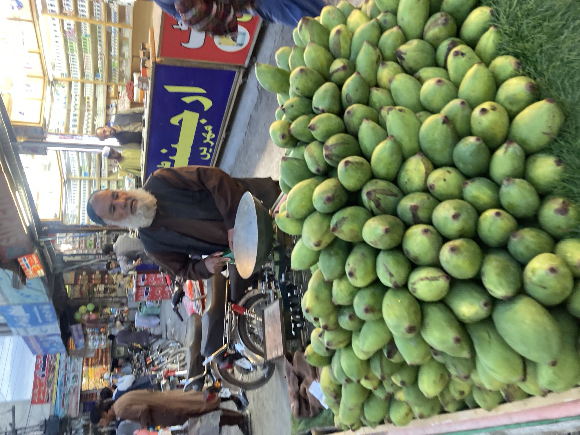 In Quetta City Tehsil, Pakistan, an outdoor vendor is selling mangoes. The vendor, an older man with a long white beard and a blue cap, wears a brown outfit. He is near a display of numerous green mangoes, piled high, and his motorbike. Other people can be seen in the background, some near storefronts.