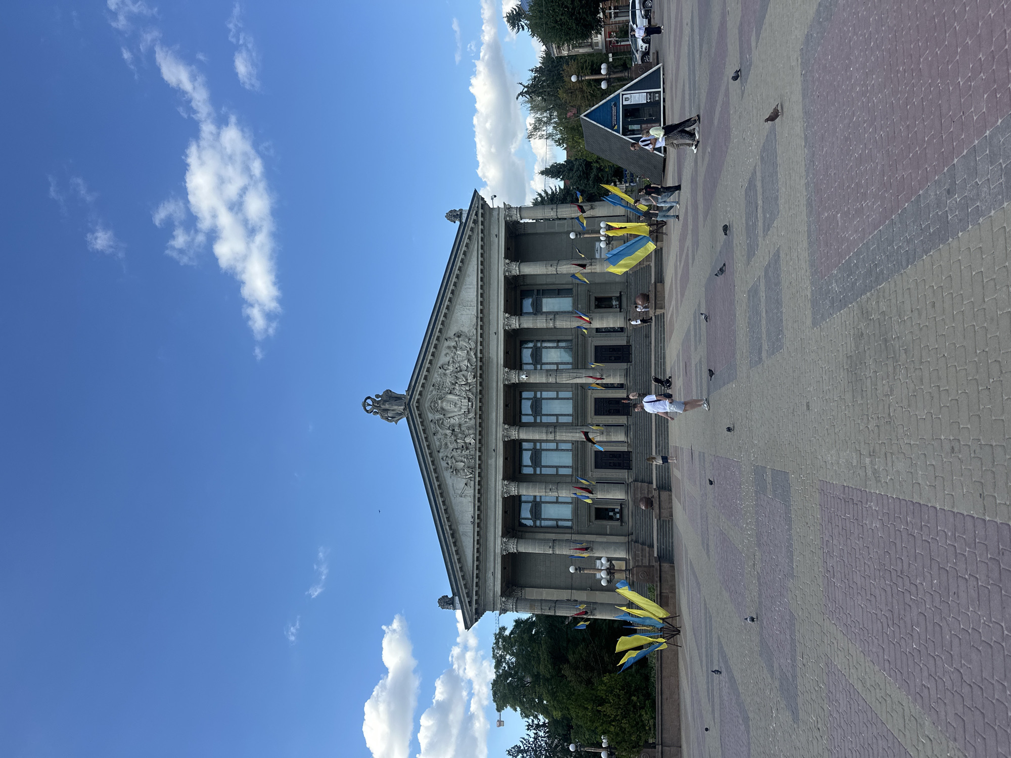 The image depicts Ternopil Drama Theatre in Ternopil, Ukraine, on a sunny day with a partially cloudy blue sky. The grand neoclassical building features a prominent portico with tall columns and a pediment adorned with sculptures. Ukrainian flags, in blue and yellow, are displayed prominently in front of the building and on flagpoles.
In the foreground, a paved plaza with a patterned brick surface is visible. Several pigeons are scattered across the plaza. A few people are walking or standing in the plaza, including a person in a white t-shirt and shorts, another person with a stroller, and a couple walking together. To the right of the theatre, a small, modern, blue, triangular structure stands near the street. Cars and trees are visible in the background. The overall scene suggests a public gathering space in front of a significant cultural landmark.