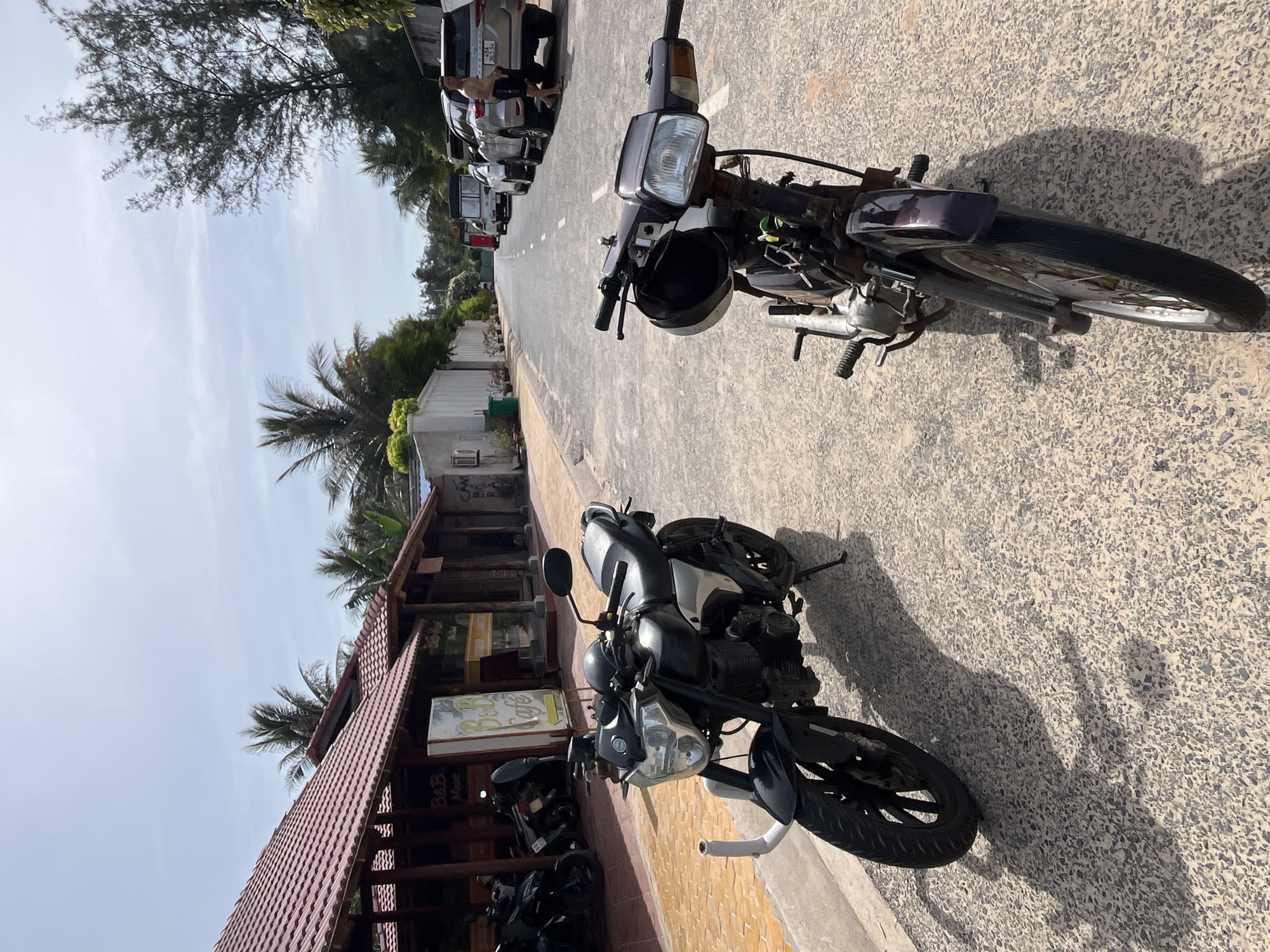 The image depicts a street scene in Phu Thuy Ward, Vietnam, on a sunny day. In the foreground, two motorcycles are parked on a paved road. The motorcycle on the left is a black and silver street bike with a prominent headlight and black wheels. The motorcycle on the right is a dark purple scooter with a visible helmet resting on its handlebars. Behind the motorcycles, a row of buildings with red-tiled roofs lines the street. One building has a sign that reads "B&B Resort" and "B&B Cafe." Further down the road, several cars are parked, including a silver SUV with a license plate visible. A man wearing a dark t-shirt and shorts stands near the silver SUV. Palm trees and other tropical foliage are scattered throughout the scene, suggesting a coastal or tropical environment. The sky is partly cloudy.
