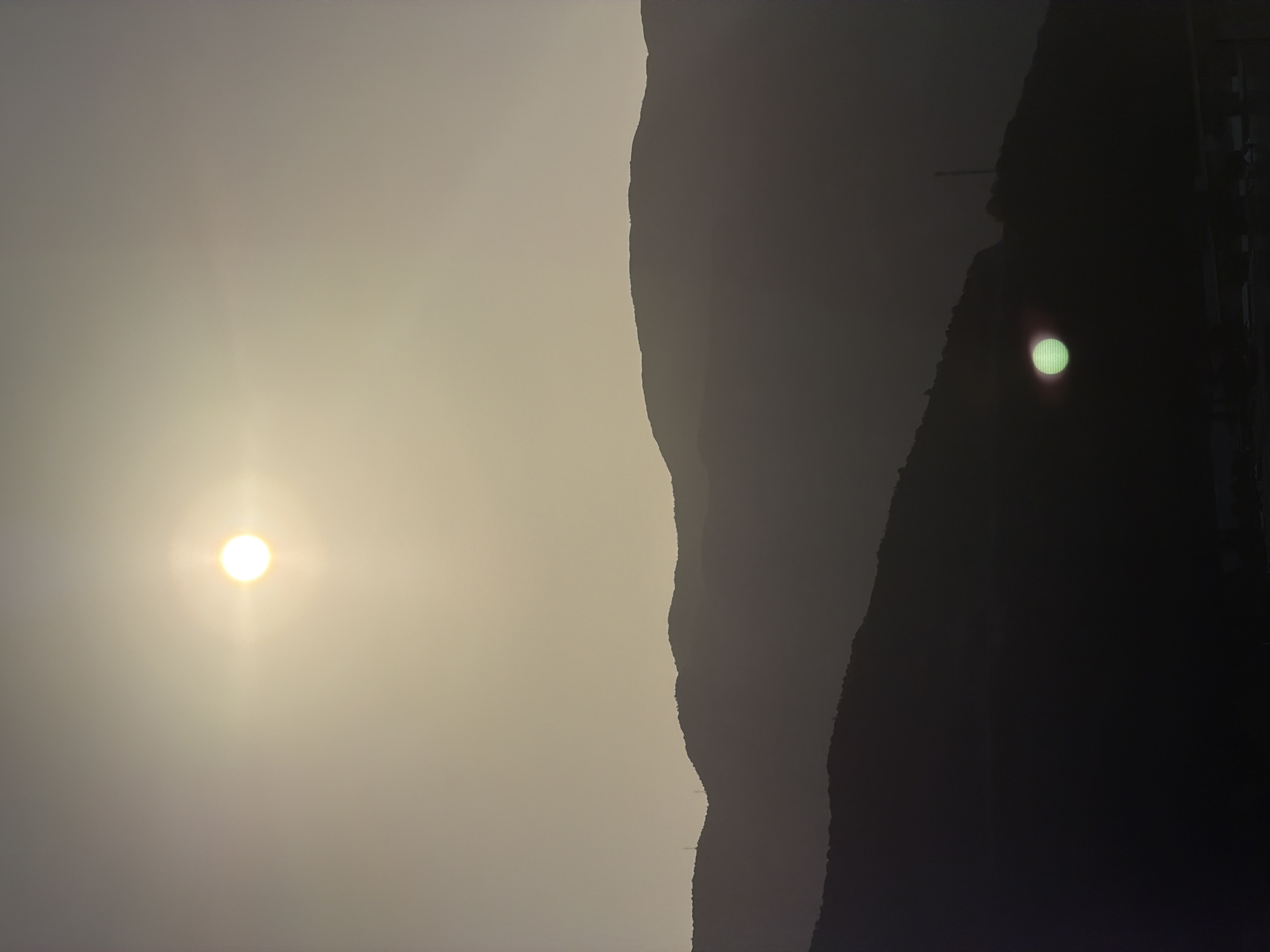 This image depicts a hazy, atmospheric view of layered mountain silhouettes against a pale sky, likely during sunset or sunrise. The sun, appearing as a bright, slightly blurred orb with a distinct lens flare, is positioned in the upper portion of the frame. Further down, a second, smaller sun-like reflection, emitting a green glow, is visible closer to the bottom of the frame, possibly a lens artifact or reflection. The mountains are dark and indistinct, with some appearing as sharp, serrated ridges. Antennas are visible on the peaks of some mountains. In the lower part of the image, the faint outlines of buildings or structures can be discerned. The overall impression is one of a serene, possibly remote, natural landscape. Without specific landmarks visible in the foreground or any identifying features, it is not possible to definitively confirm the location in Budva, Montenegro.
