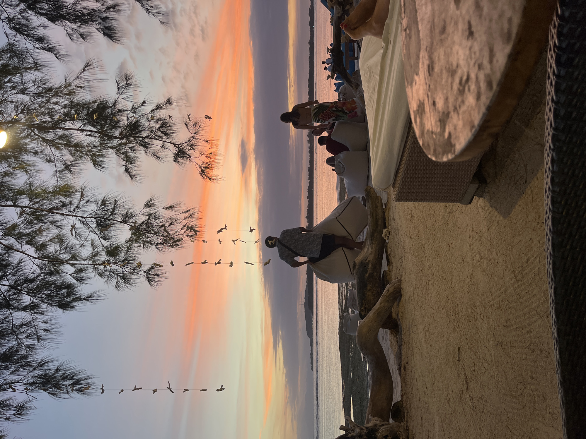 The image captures a waterfront scene at dusk, possibly in an outdoor seating area. To the left, a tree with dense foliage frames the sky. The sky is a mix of orange, pink, and grey, suggesting a sunset or sunrise. A string of bird-shaped decorations hangs in the air. Several individuals are visible: one is standing by a white sofa with patterned clothing, and others are in the background near the water, with a few visible on a boat. A round wooden table is partially visible on the right, along with a woven structure and a textured wall. The base of the seating area is constructed with driftwood elements. The location is Unknown, Indonesia.