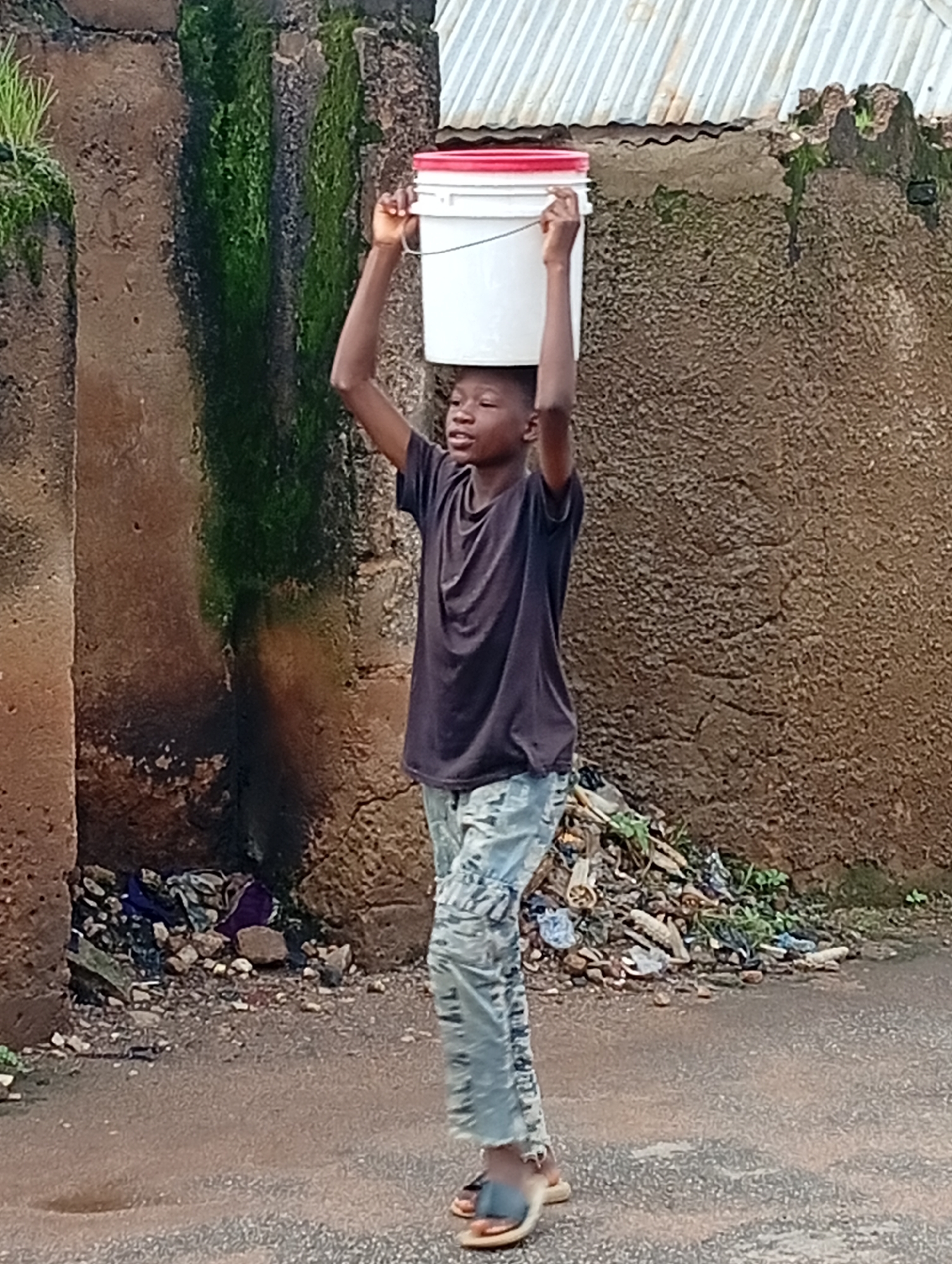 A young boy walks with a white bucket on his head, balancing it with his right hand. He is wearing a dark t-shirt and camouflage-patterned trousers. The scene appears to be outdoors, possibly on a street or dirt path, with a textured, brown wall behind him. There is some debris and greenery near the wall. The environment suggests a casual, possibly residential area. The image is taken in a medium shot, focusing on the boy and his action. The lighting is natural, indicating daytime.