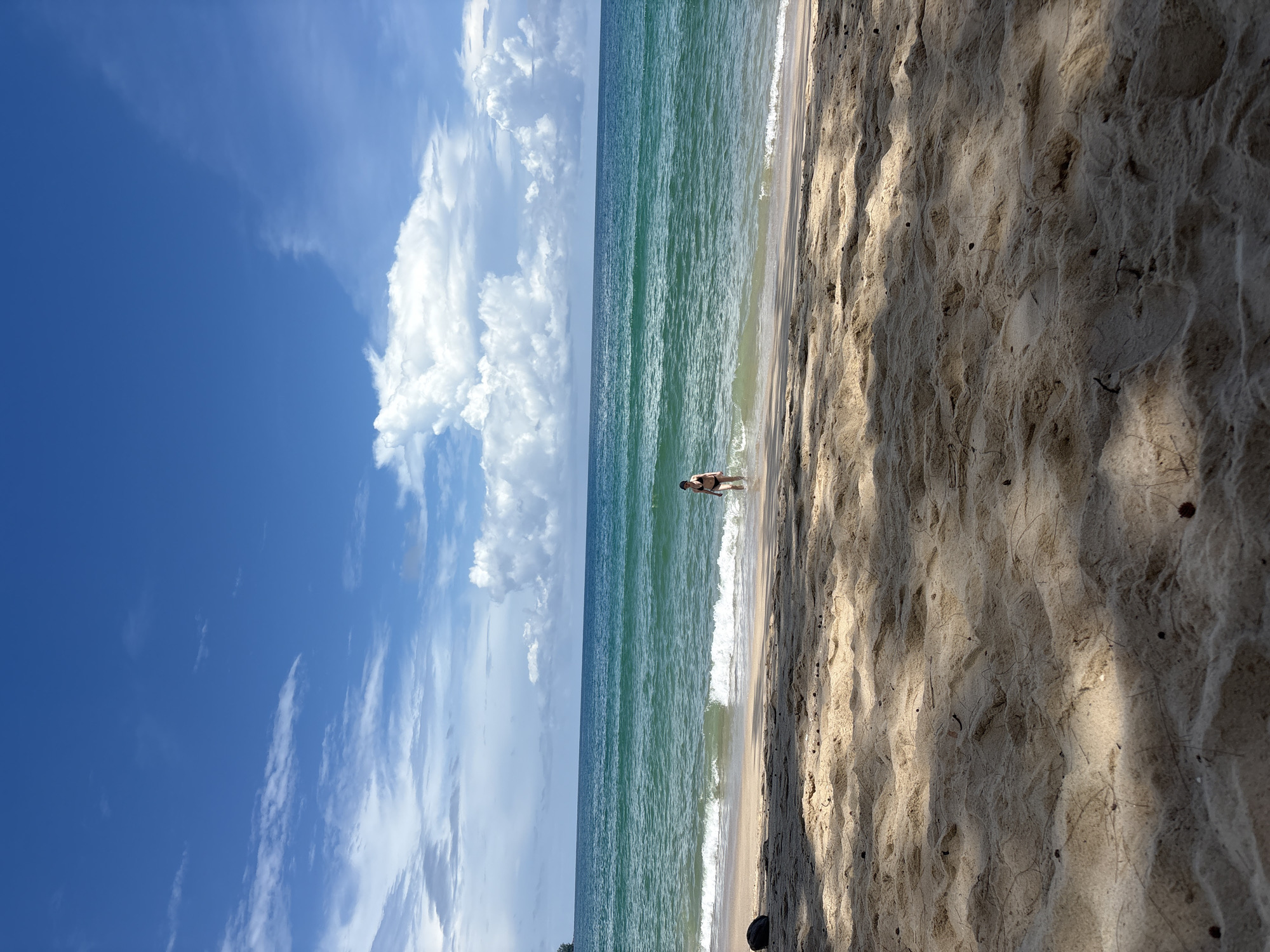 In Choeng Thale, Thailand, on a sunny day, a person wearing a black bikini is standing in the shallow water at the edge of the ocean on a sandy beach. The water is a clear turquoise, with gentle waves lapping at the shore. The sand is light brown and appears to be untouched, with shadows cast by unseen trees. The sky above is a vibrant blue, with scattered white clouds.