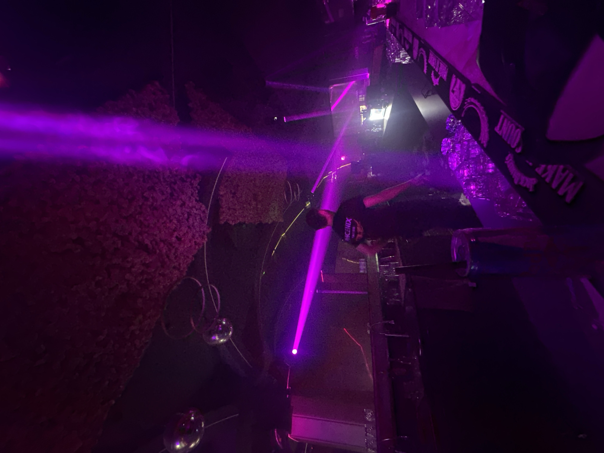 The image captures the interior of a dimly lit nightclub or bar, saturated with magenta and purple lighting. The location is in Sliven, Bulgaria, as indicated by the word "SLIVEN" visible on the back of a staff member's shirt.
In the mid-ground, a male individual is positioned behind a long bar counter, facing away from the viewer. He wears a dark, short-sleeved t-shirt with a white logo on the back that appears to read "PHOENIX" or similar, with "SLIVEN" printed beneath it. His arms are slightly bent, suggesting active engagement with the bar area.
The bar counter extends across the lower portion of the frame. It is dark-colored and features several beverage taps. Numerous clear drinking glasses are stacked neatly on the counter. In the lower right corner, a silver and blue Red Bull can with a black straw is present on the counter, adjacent to a black rubber bar mat that displays white brand logos, including "Jack Daniel's" and the phrase "MAKE YOUR COUNT." Behind the bar, sections of the wall or shelving are illuminated with a bright white light.
The ceiling is dark, with portions covered in a textured, possibly artificial foliage or acoustic material, which is bathed in purple light. Multiple spherical, mirrored disco balls are suspended from the ceiling, reflecting the vibrant purple light. Thin, curved metallic structures are also visible on the ceiling, likely part of the lighting rig. Several intense, focused beams of purple light cut through the hazy air, originating from various points and creating dynamic patterns throughout the space and across the surfaces of the bar.