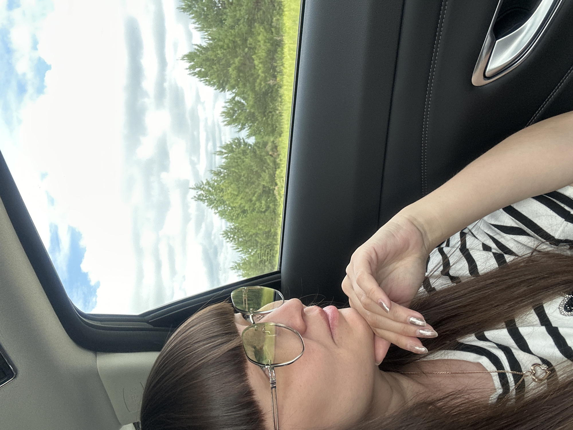 In the city of Dokshitsa, Russia, a woman with long, dark brown hair and bangs is seated inside a vehicle, looking out the window. She is wearing large, round, tinted sunglasses and a striped shirt. Her left hand is resting on her chin, with her fingers displaying light-colored, glittery nail polish. A delicate gold necklace with a heart-shaped pendant is visible around her neck. The interior of the car features black leather upholstery. Outside the window, a landscape of green trees and a cloudy sky is visible.