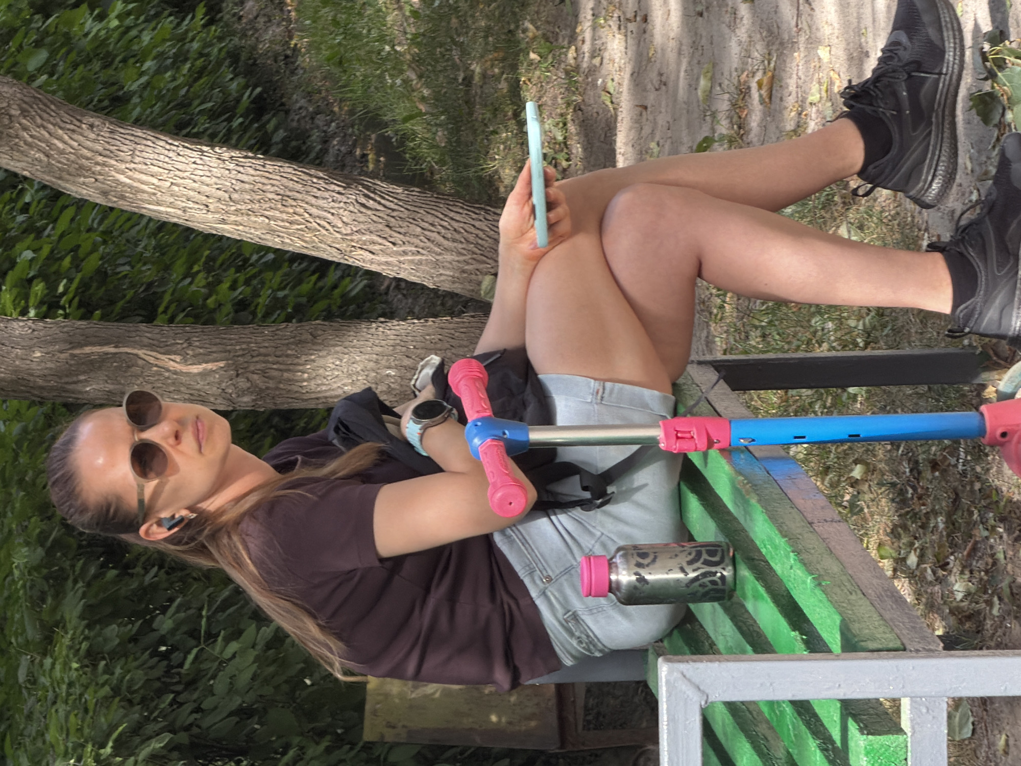 A woman with brown hair pulled back in a ponytail, wearing sunglasses and earbuds, sits on a green and gray slatted park bench. She is wearing a dark maroon t-shirt, light blue denim shorts, black socks, and black athletic shoes. A blue and pink scooter is positioned in front of her. To her left on the bench is a metallic water bottle with a pink lid. In her right hand, she holds a light blue smartphone. The background consists of trees, green foliage, and dry leaves on the ground.