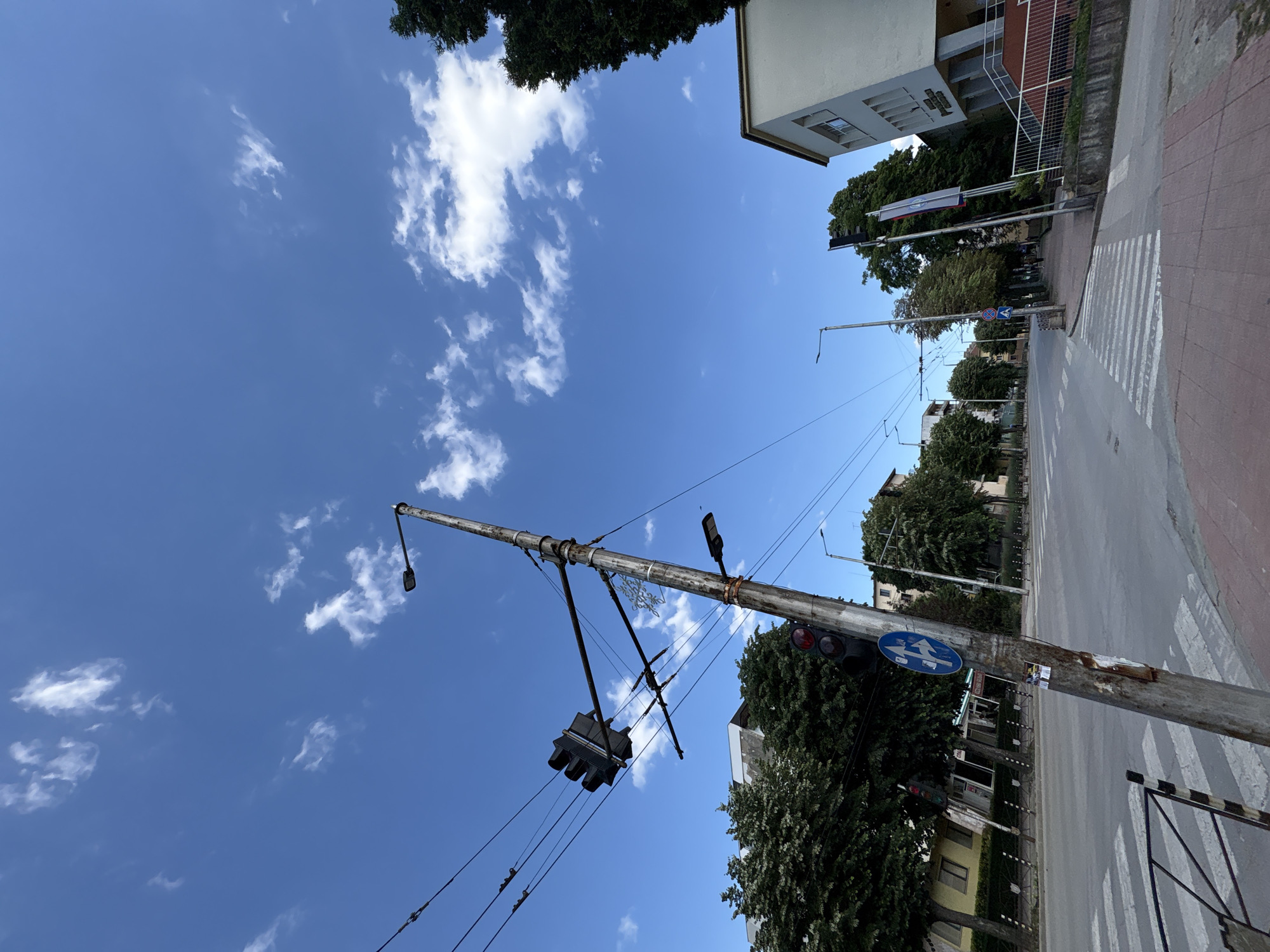 This is a street-level, wide-angle photograph taken in the city of Vratsa, Bulgaria. The perspective is looking upwards towards a bright blue sky with scattered white clouds. In the foreground, a grey, weathered utility pole leans to the left, supporting traffic lights and overhead power lines. The traffic lights are mounted at different heights and angles. One set of traffic lights appears to be red. A blue circular traffic sign with white arrows indicating a turn is attached to the pole, along with some stickers. The pole is positioned near a pedestrian crossing marked with white stripes on the asphalt road.
To the left of the pole, a street stretches into the distance, lined with trees and buildings. The buildings appear to be residential or commercial, with windows and architectural details. The street has power lines running above it, supported by other poles.
To the right of the utility pole, a building with a white facade and grey trim is visible. This building has windows and an entrance with stairs leading up. There is a low concrete wall in front of this building. Green trees are present throughout the scene, some tall and dense, others smaller and interspersed with the buildings. The pavement in the foreground is paved with reddish-brown bricks. The overall impression is that of a typical urban street scene on a clear day.